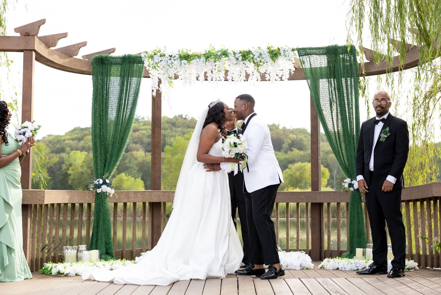 Bride and groom share first kiss at outdoor wedding ceremony under green draped arch on wooden deck.