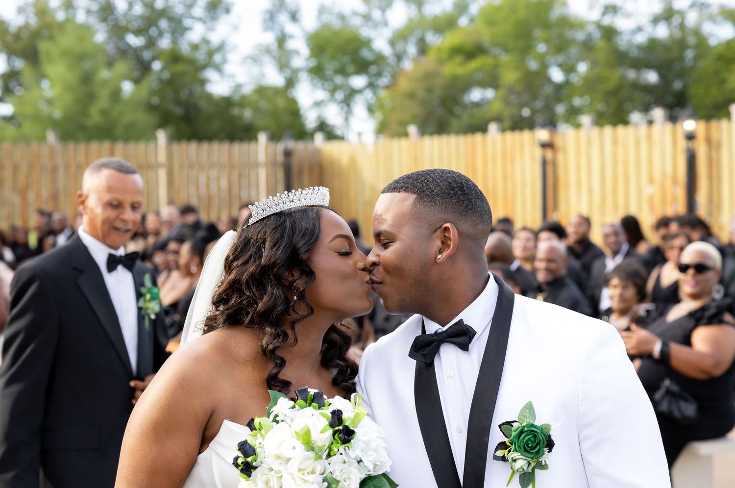 Bride and groom share a kiss at outdoor wedding ceremony, bride wearing tiara and white gown, groom in white tuxedo jacket.