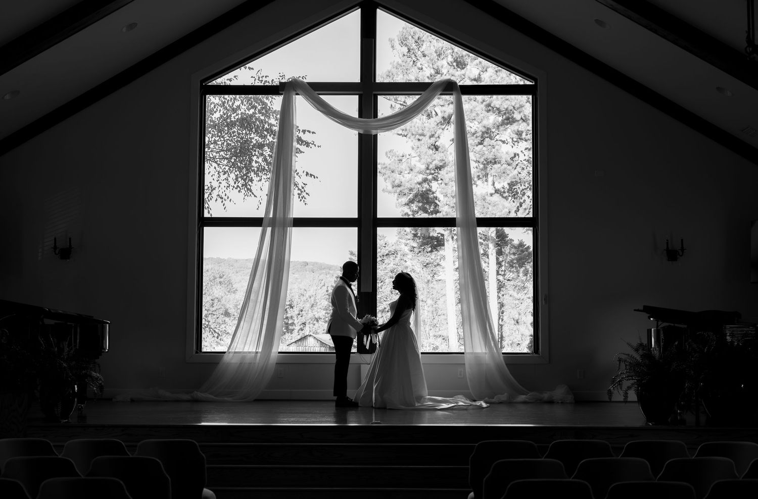 Black and white silhouette of a bride and groom holding hands at the altar of a chapel with large windows.