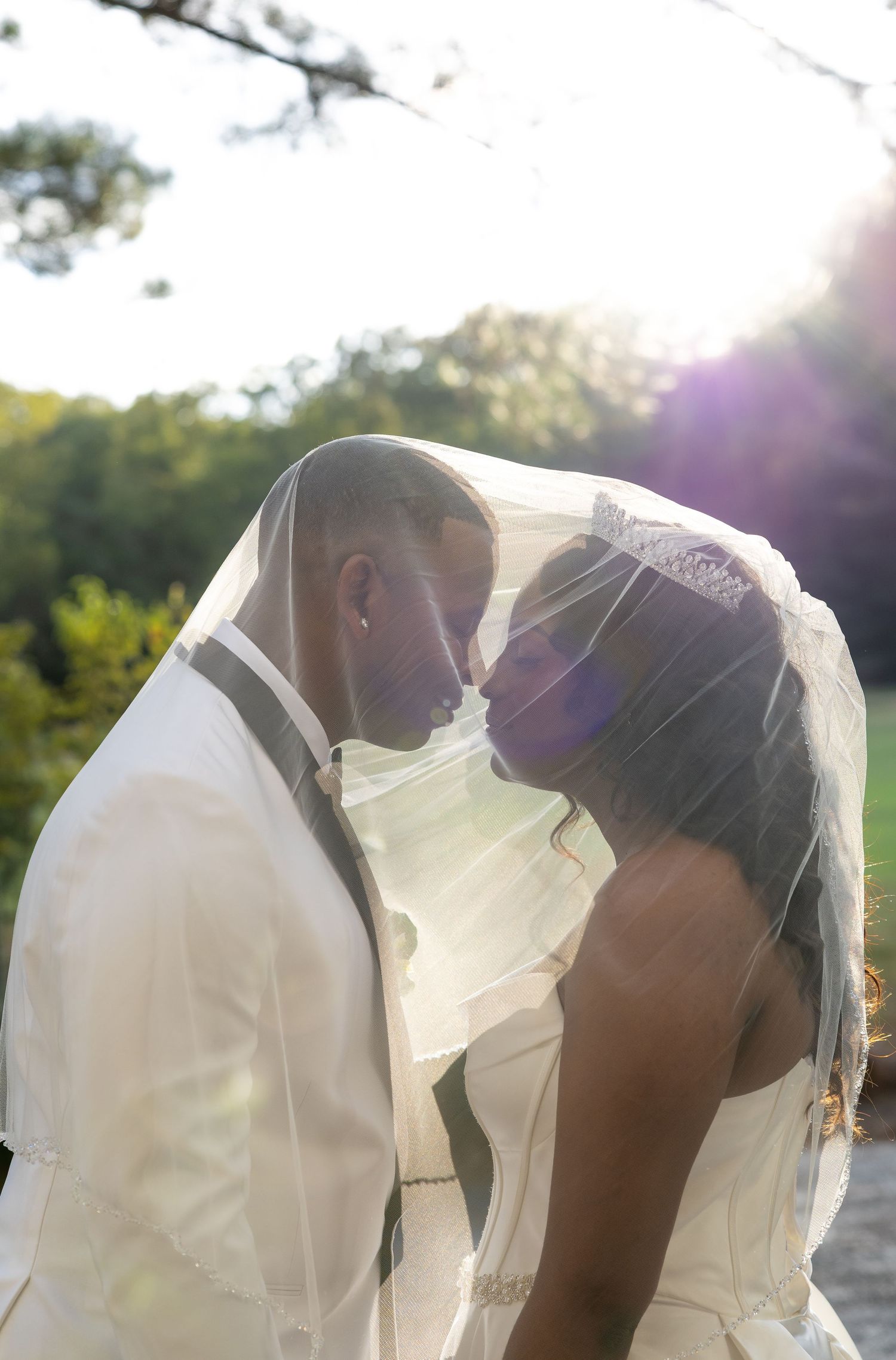 Bride and groom sharing a romantic kiss under a veil in golden sunlight on their wedding day.