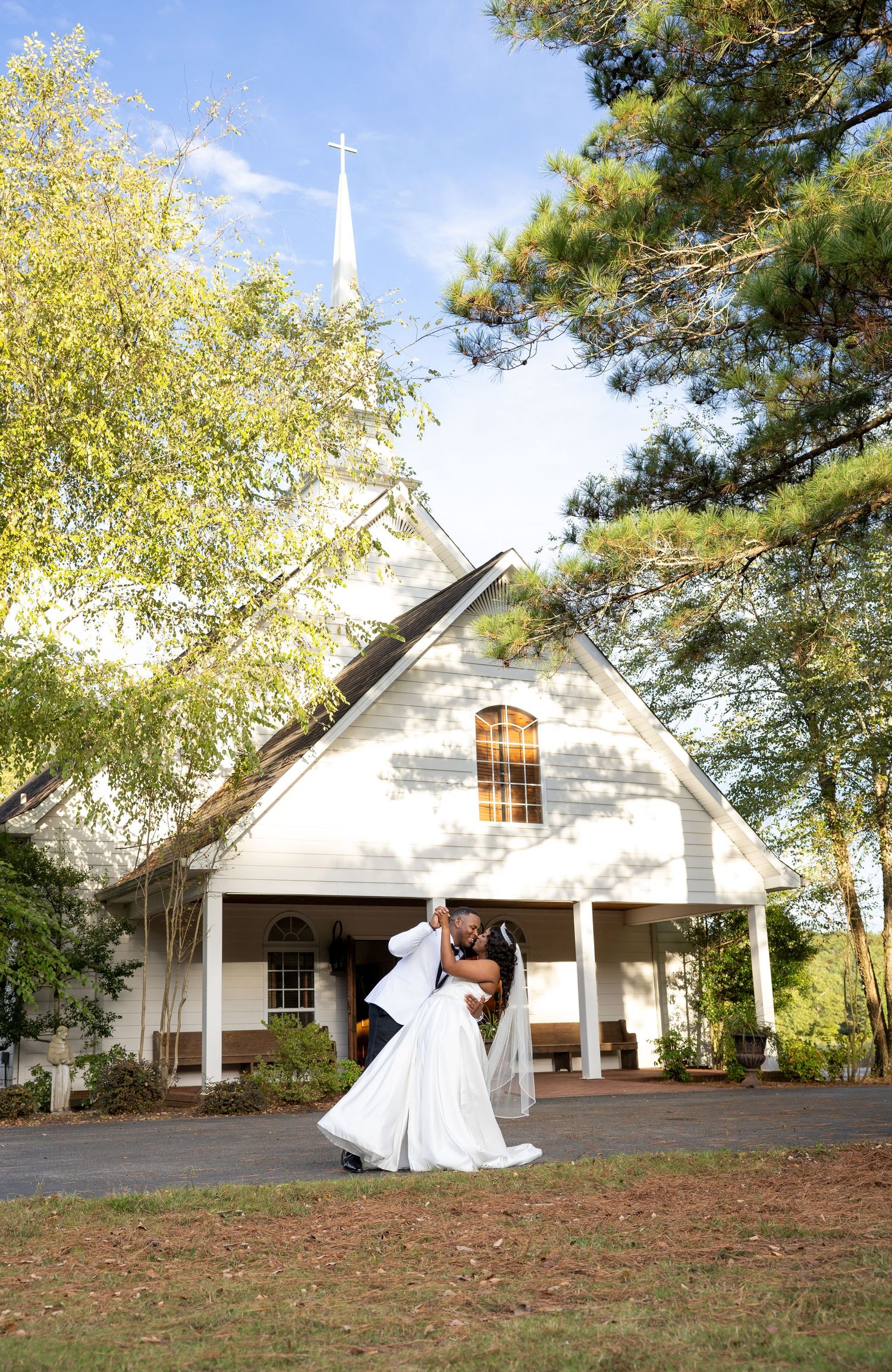 Newlywed couple sharing a romantic dip kiss outside a white chapel church surrounded by trees on their wedding day.