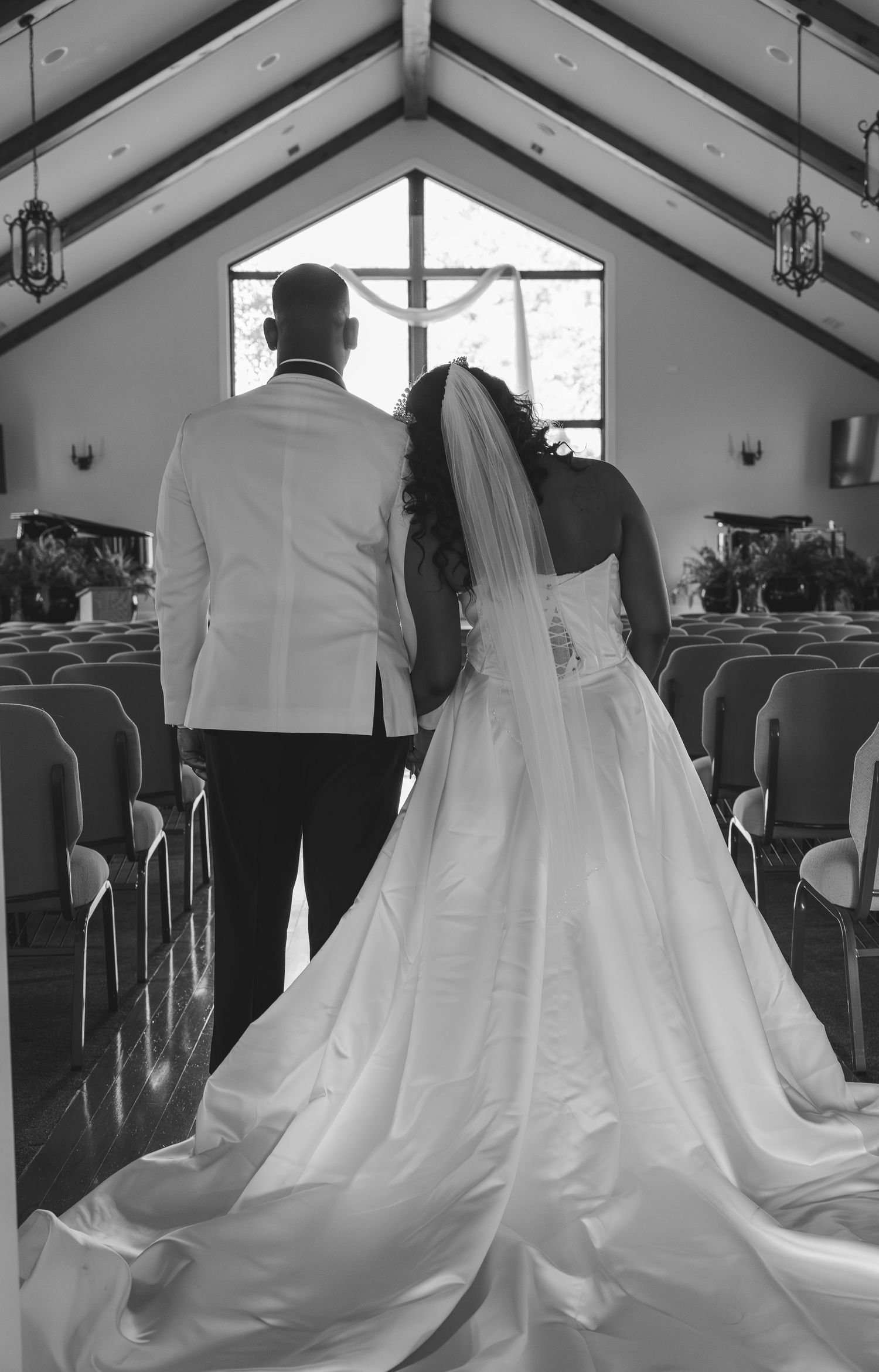Black and white photo of bride and groom walking down church aisle toward altar window on wedding day.