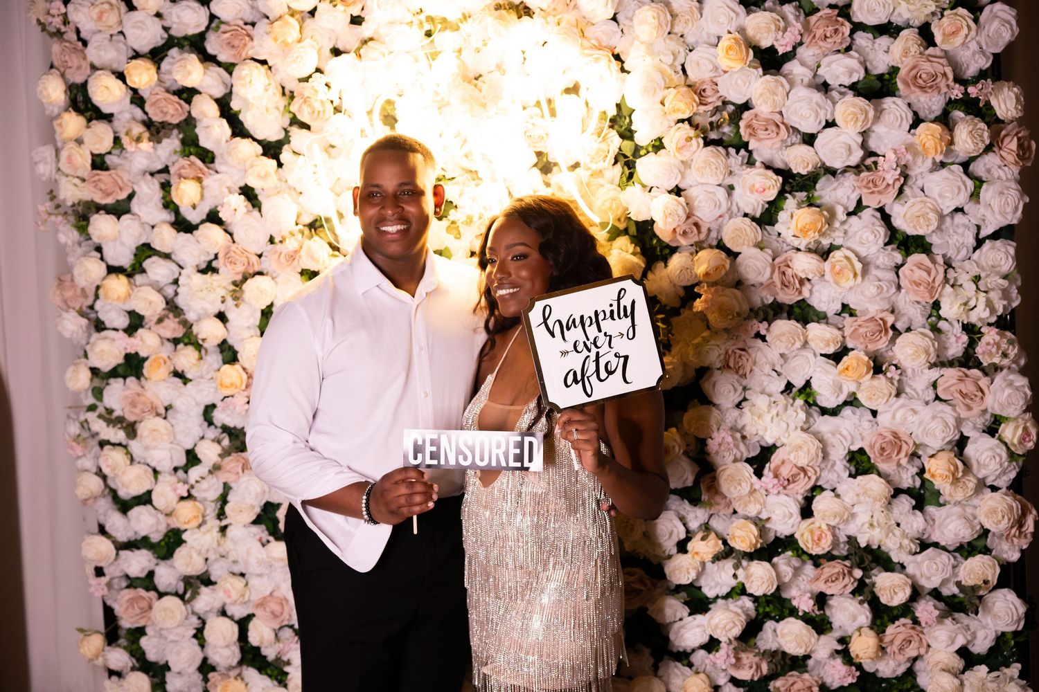 Couple posing at floral photo booth with censored signs at wedding reception.