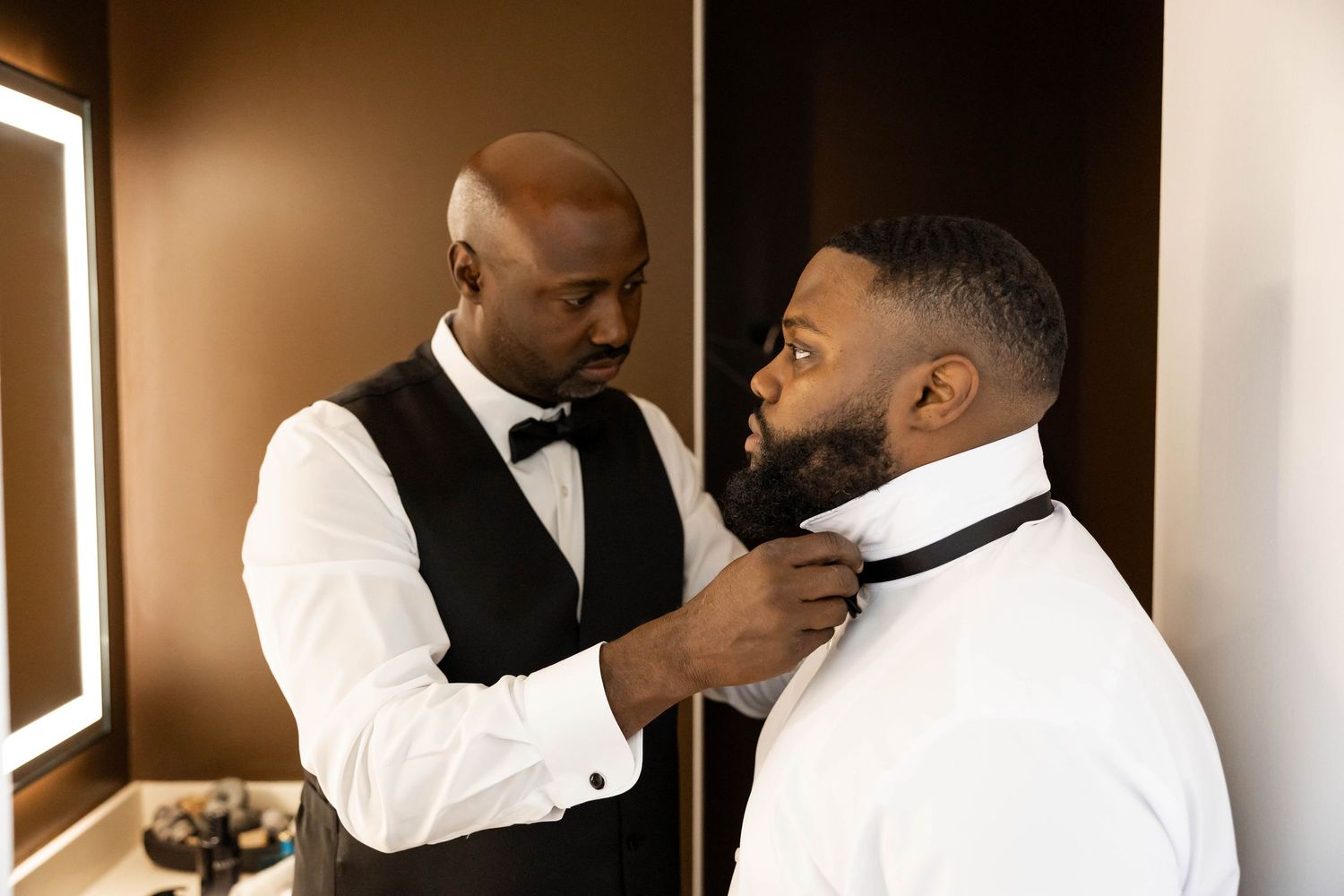 A groomsman adjusts the bow tie of a groom in a white shirt during wedding preparations.