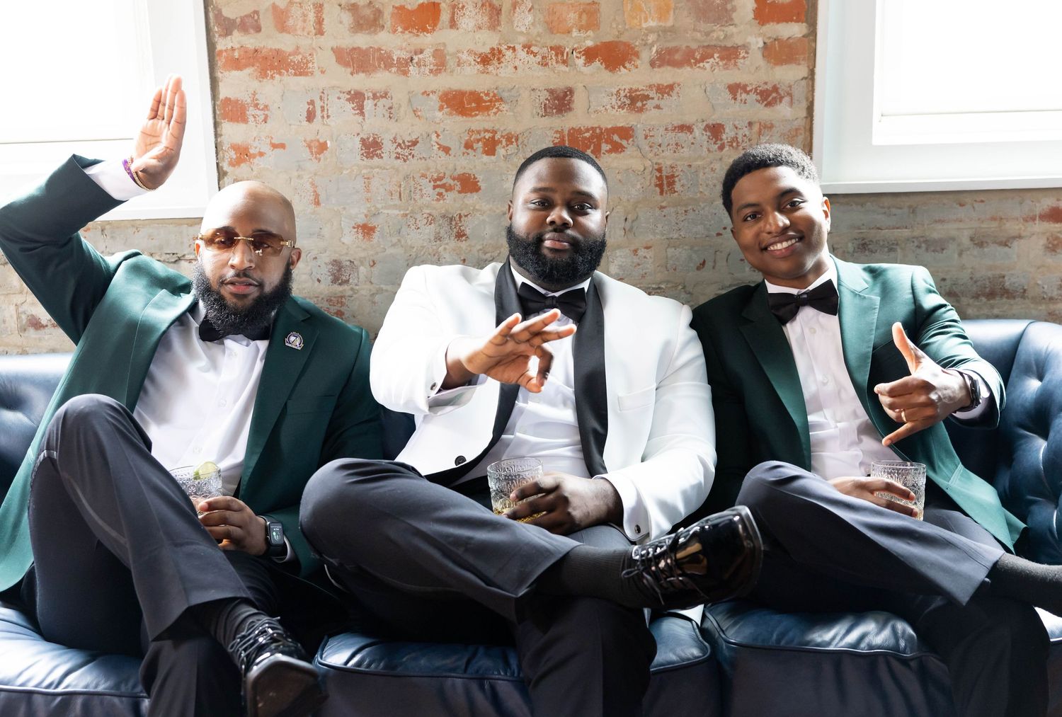 Three men in elegant green and white tuxedos pose confidently on a blue velvet sofa in a stylish venue.