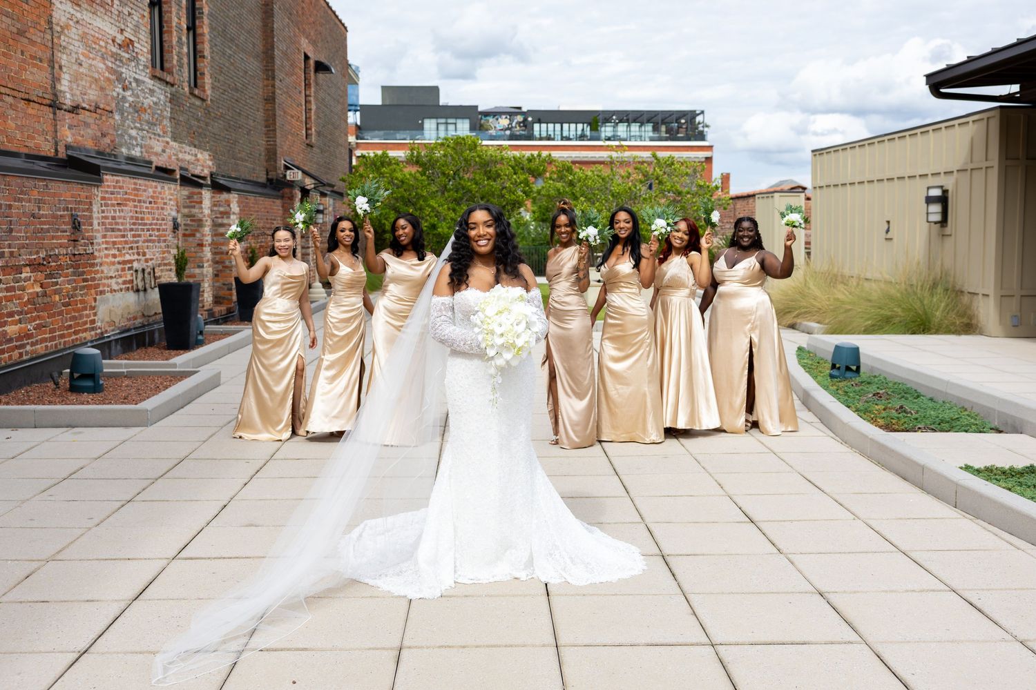 Bride in white gown with bridesmaids in gold satin dresses posing outdoors on urban patio for wedding photo.