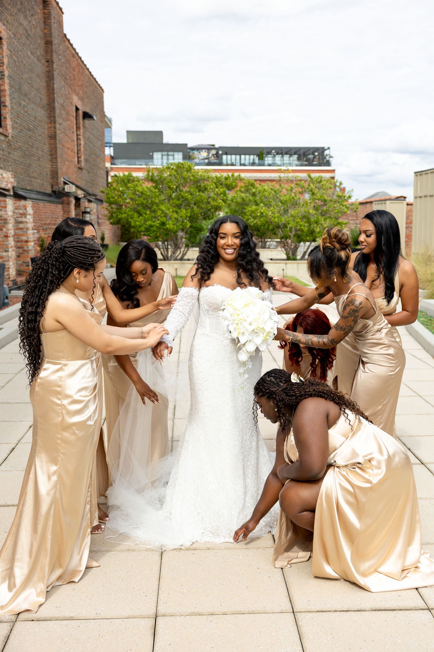 Bride in white lace gown holds bouquet while bridesmaids in gold satin dresses assist her on an outdoor rooftop.