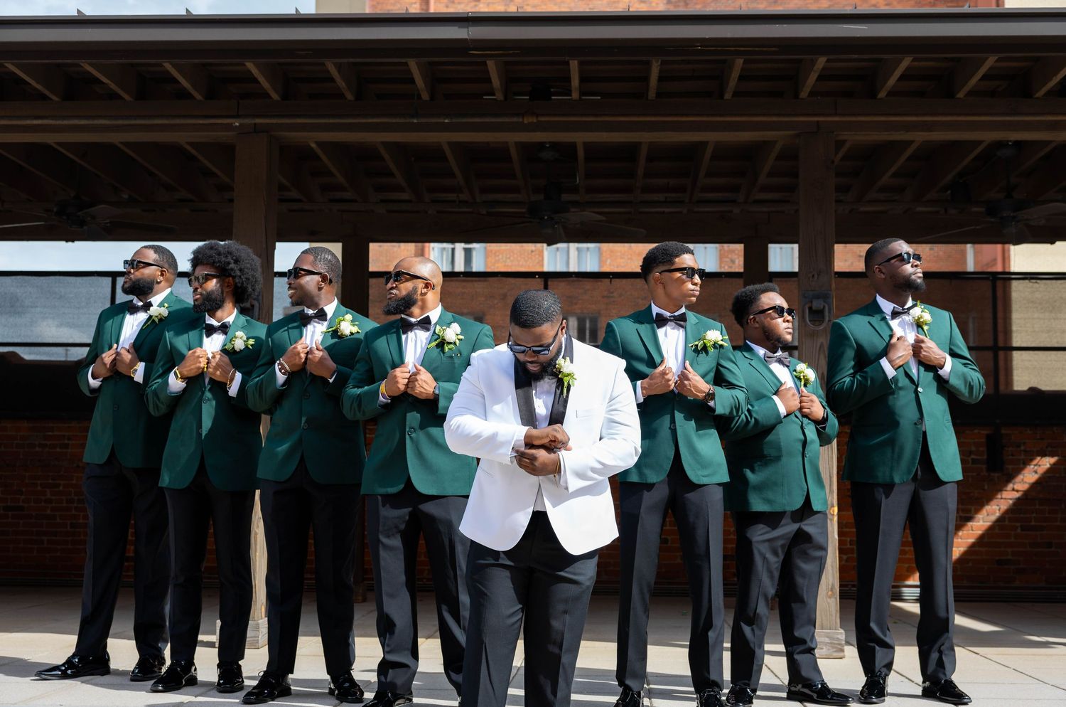 Groom in white tuxedo jacket poses with groomsmen in matching green blazers and black pants at outdoor wedding.