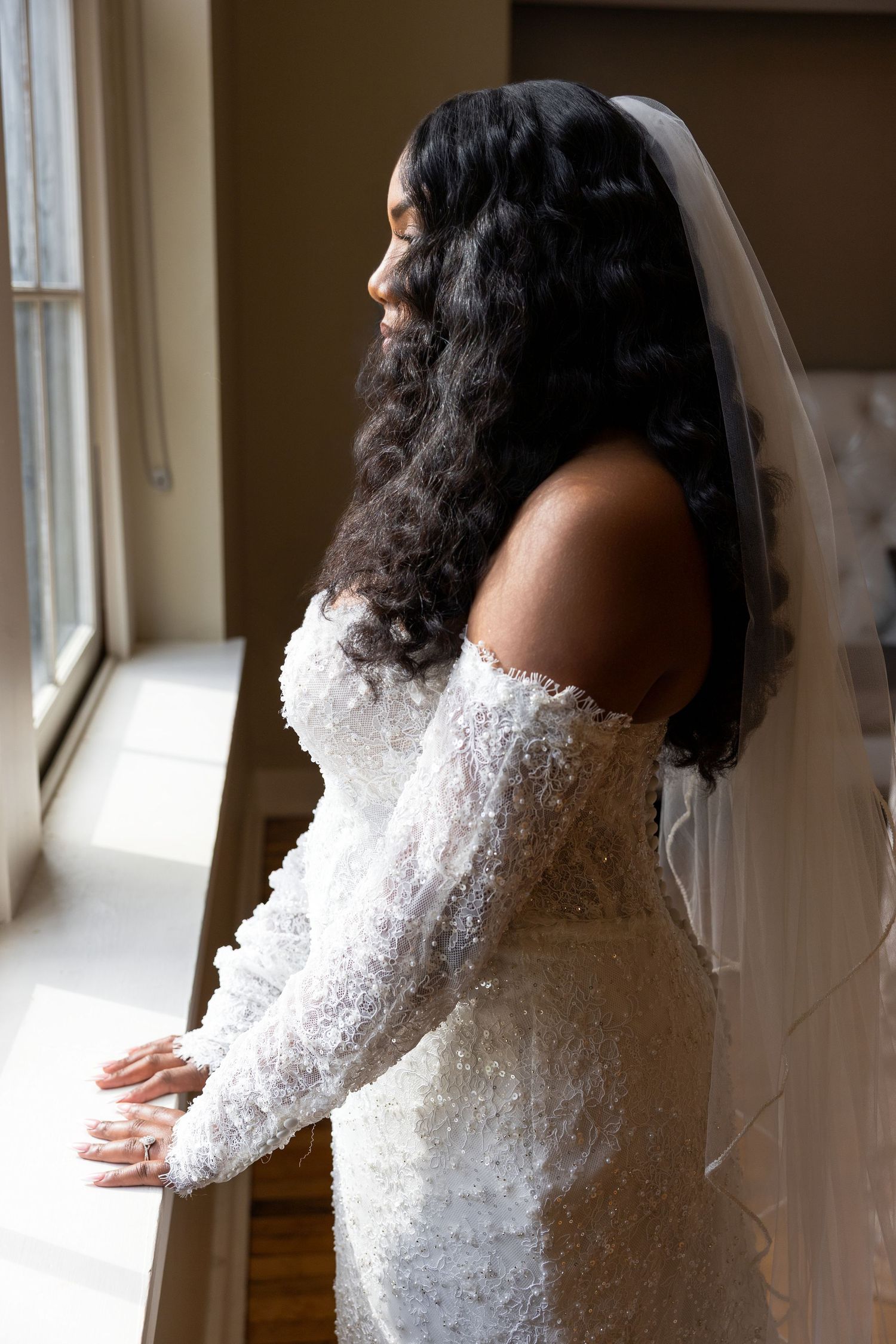 Black bride in beaded lace off-shoulder gown and veil gazes out window in elegant bridal portrait.
