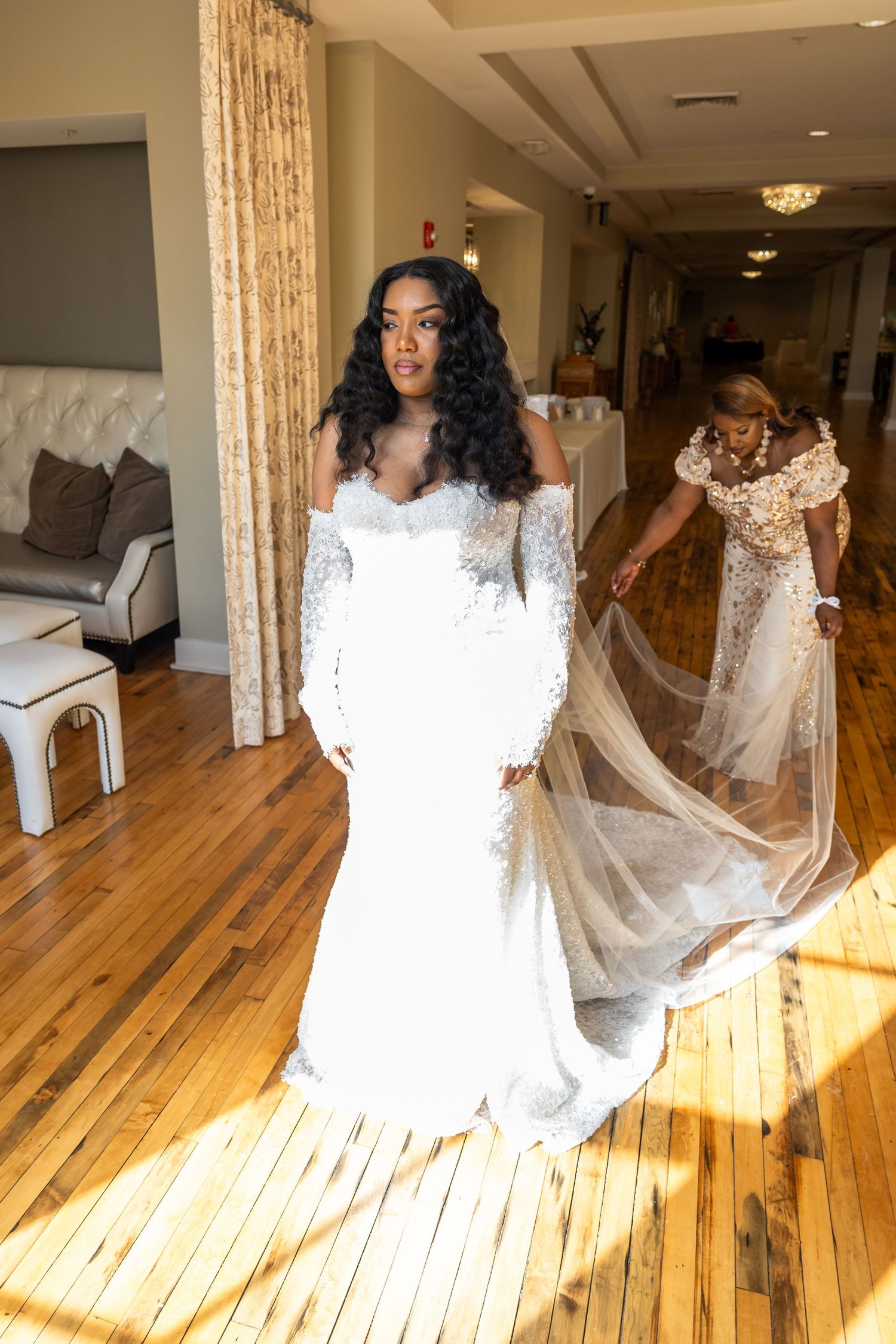 Bride in off-shoulder white lace gown with veil being spread by bridesmaid on hardwood floor.