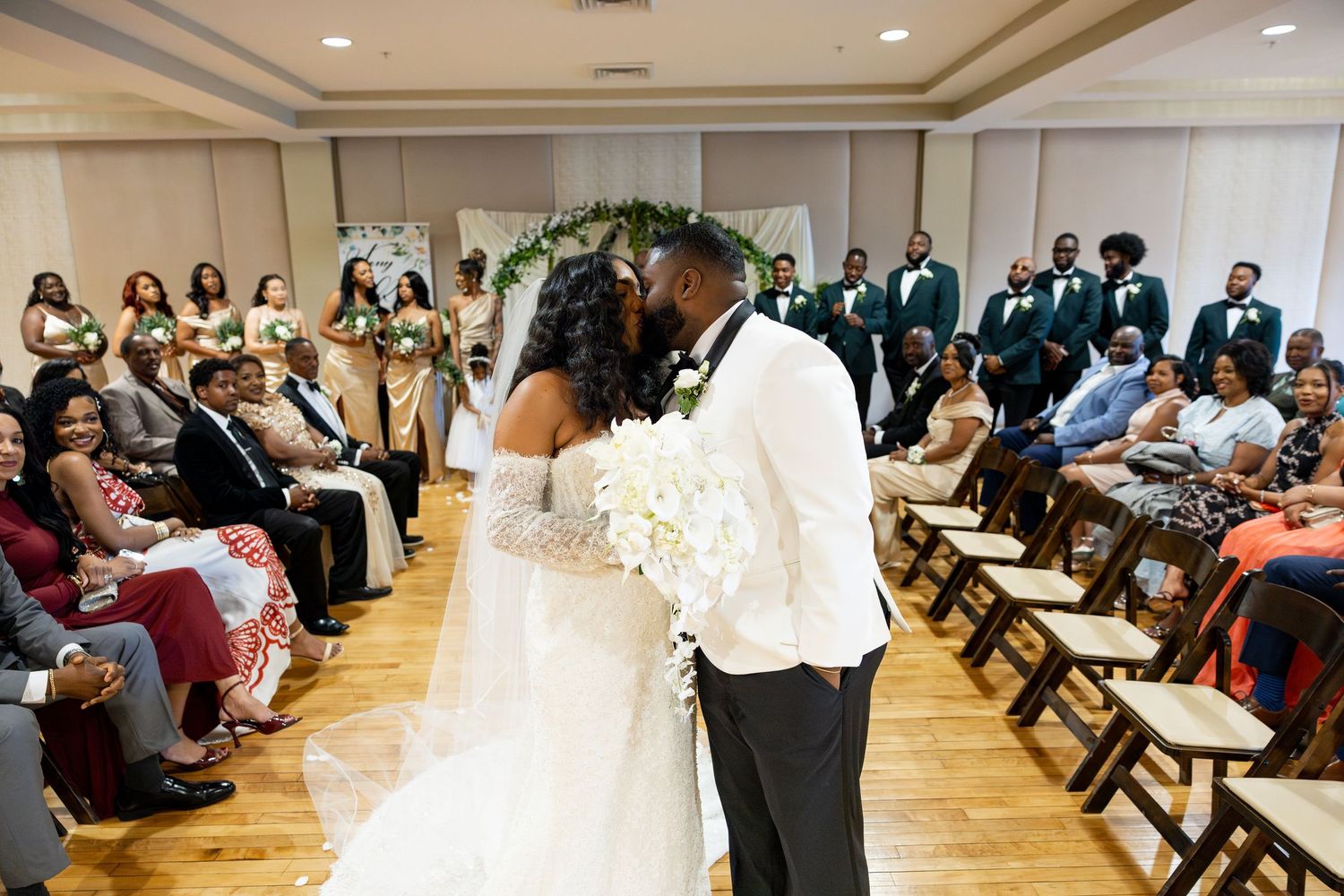 Bride and groom share first kiss at indoor wedding ceremony with bridesmaids and groomsmen standing in elegant venue.