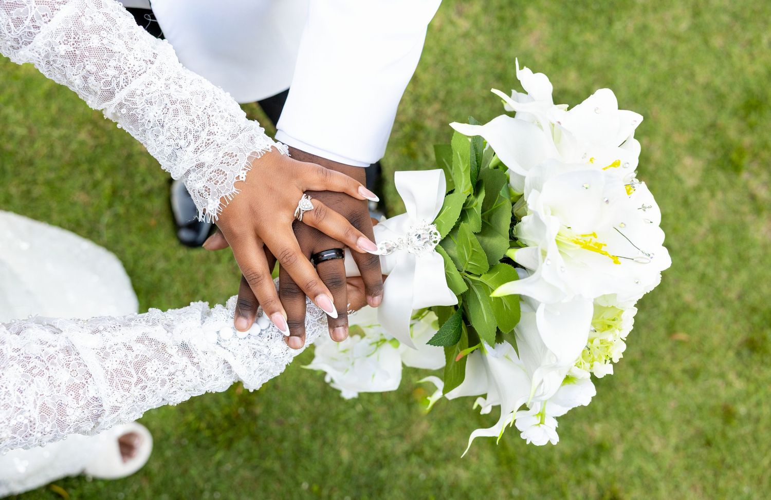 Aerial view of newlyweds holding hands with a white floral bouquet on green grass at a wedding ceremony.