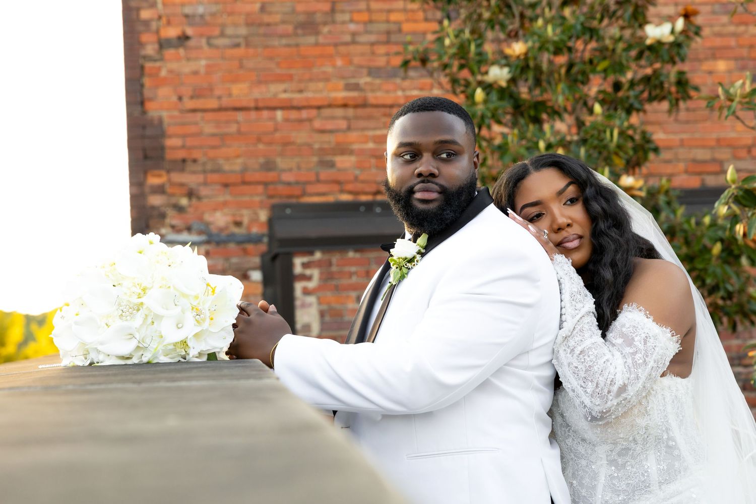 Bride and groom in elegant white wedding attire pose outdoors with a white floral bouquet against a brick wall backdrop.