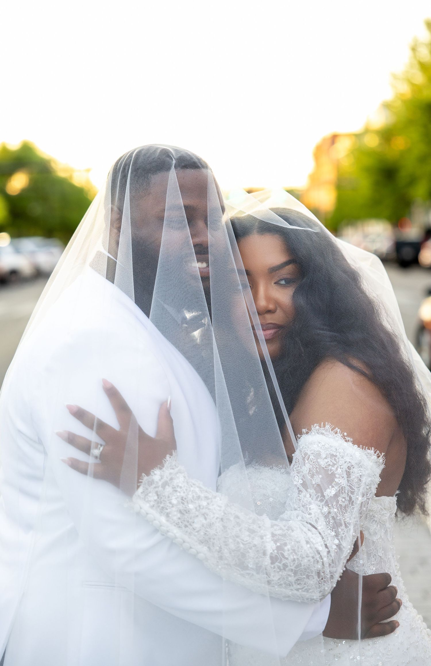 A bride and groom share a romantic embrace under a white wedding veil outdoors during golden hour.