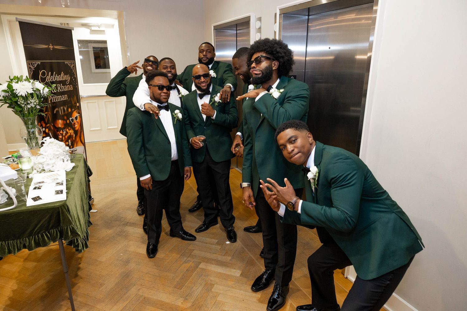 Groomsmen in emerald green tuxedos posing playfully together at a wedding venue with hardwood floors.
