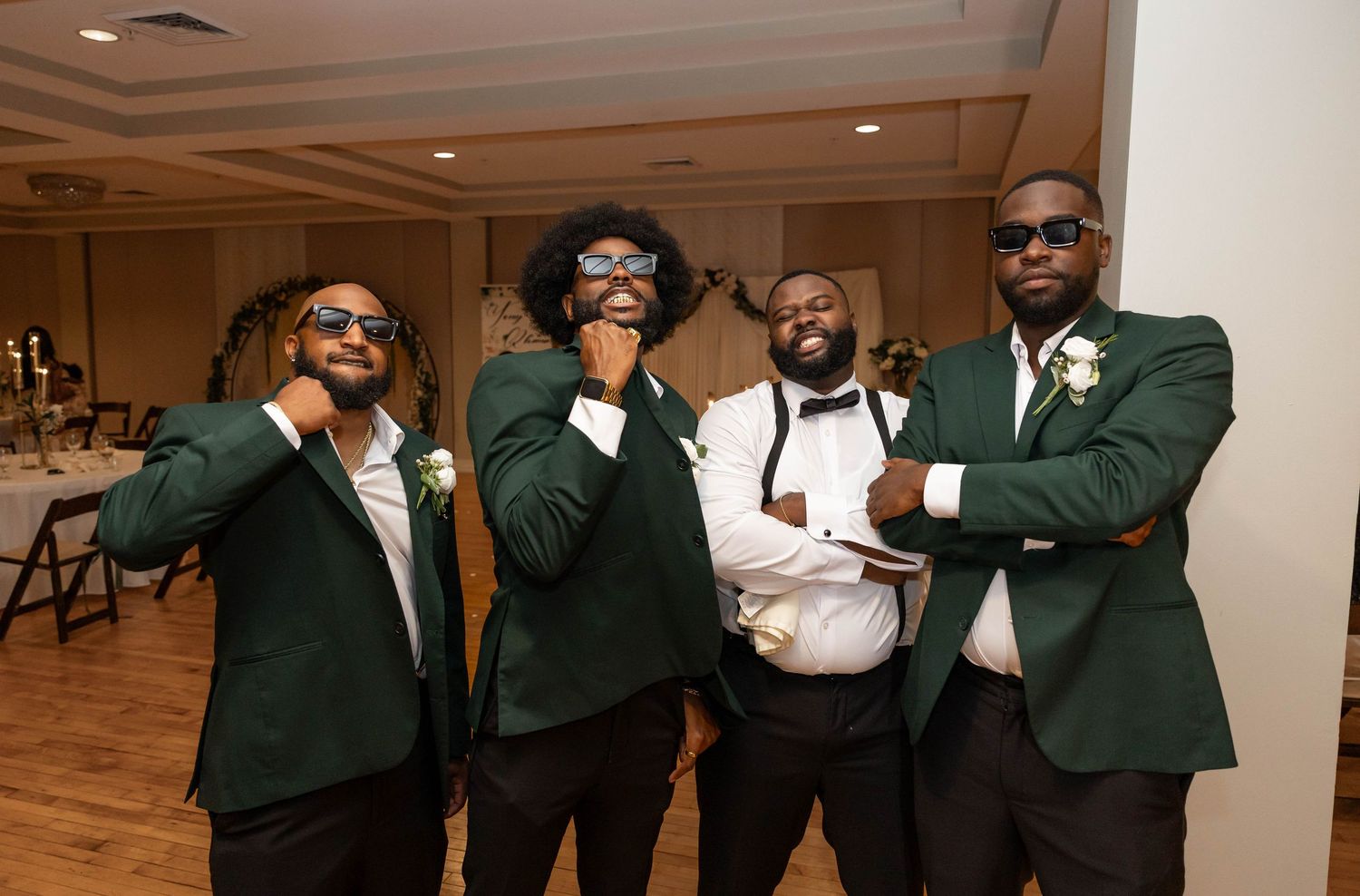 Four groomsmen in dark green suits with boutonnieres posing playfully at a wedding reception venue.
