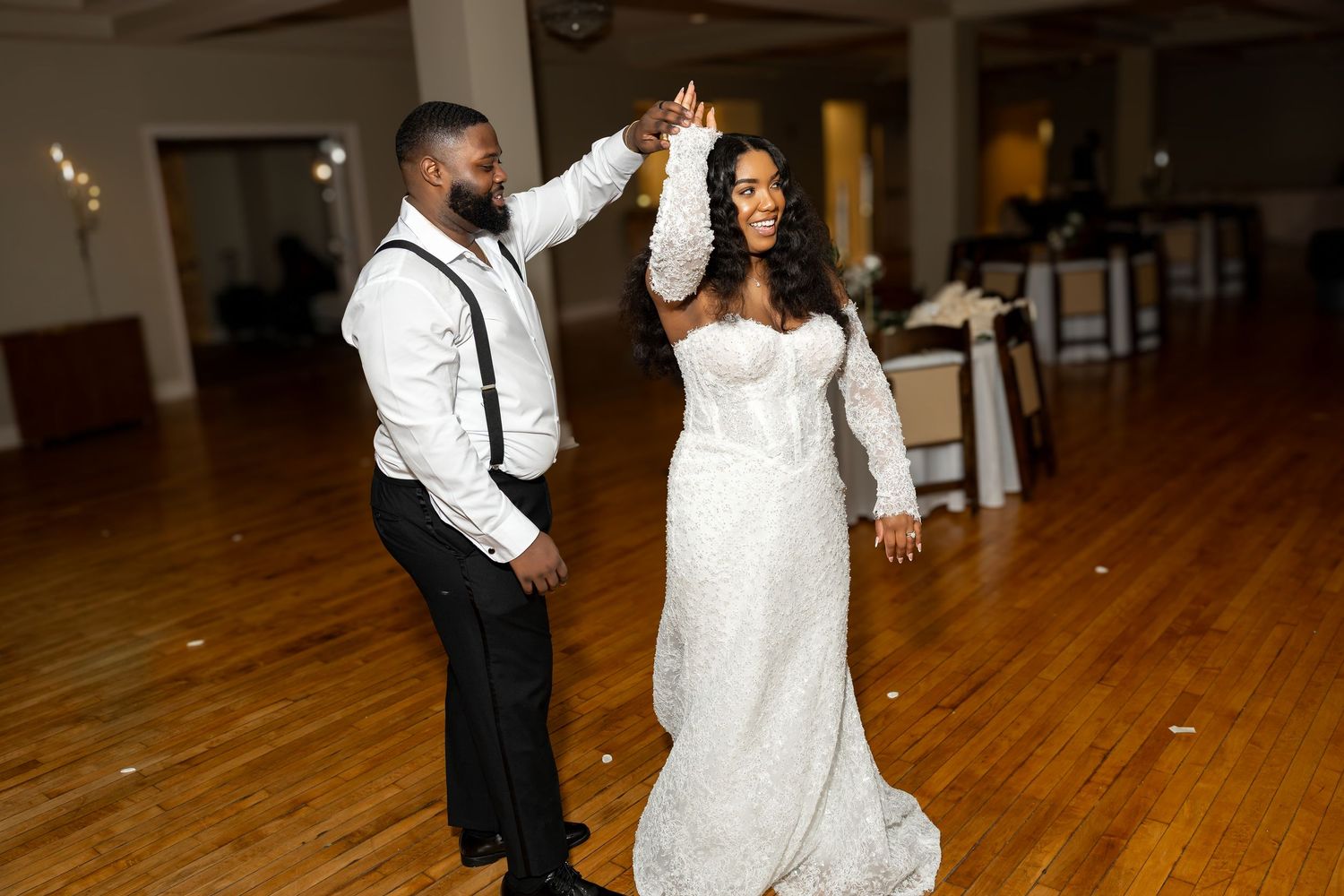 Bride and groom share first dance, groom twirling bride in white lace wedding gown on elegant ballroom floor.