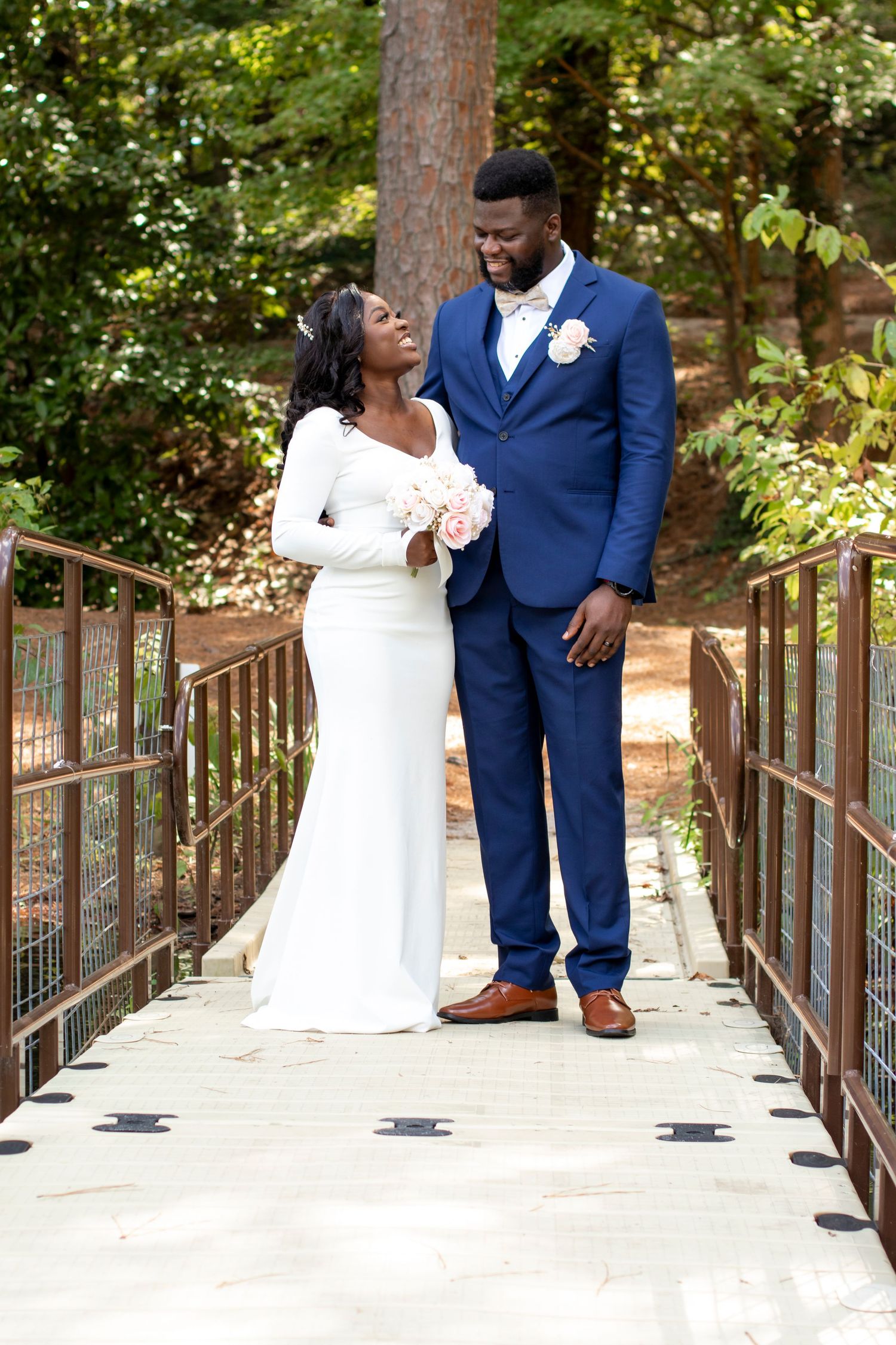A beautiful wedding couple stands together on a wooden bridge surrounded by lush greenery in an outdoor setting.