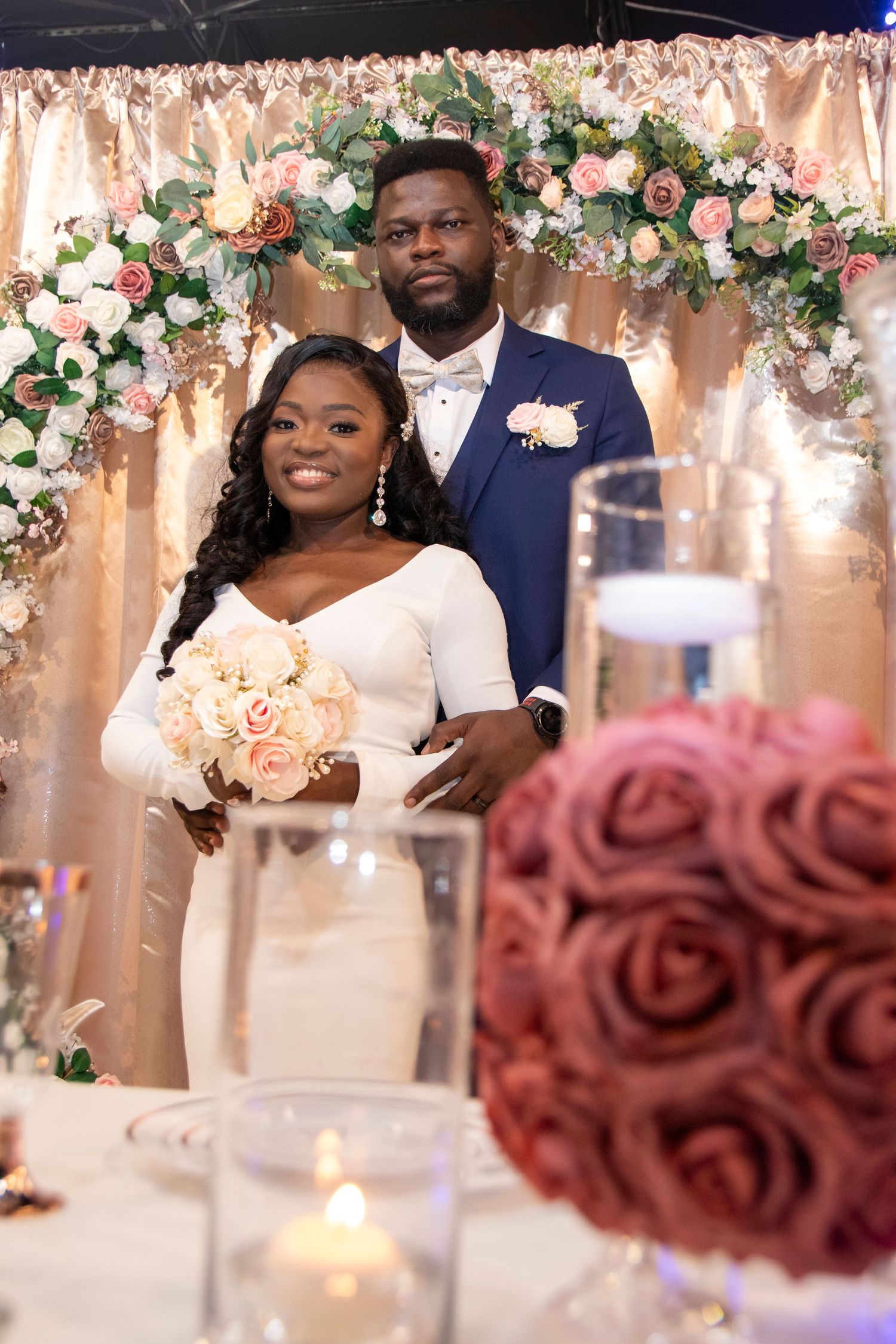 A wedding couple poses beneath an elegant floral arch with pink and white roses and candlelit table decor.