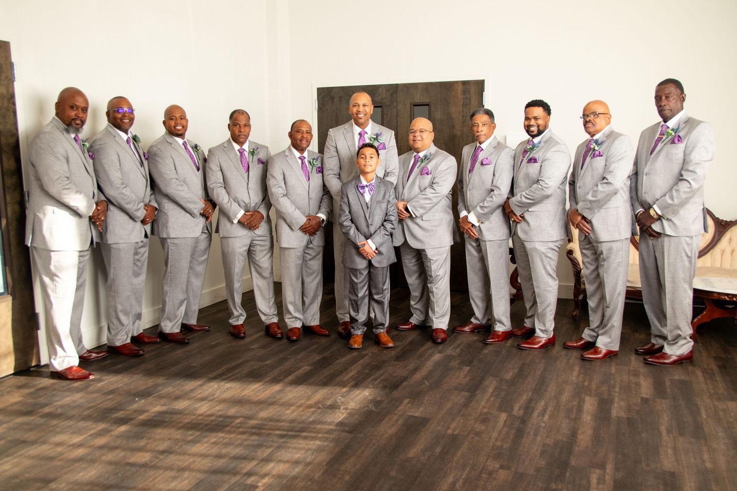 Wedding party wearing matching light gray suits and brown shoes pose together on wooden floor in formal group photo.