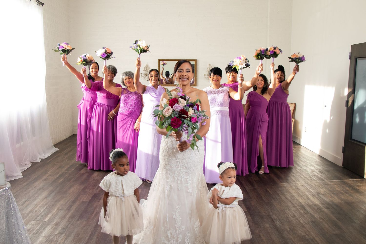 A joyful wedding party in a room with wooden floors, featuring bridesmaids in purple dresses and flower children.