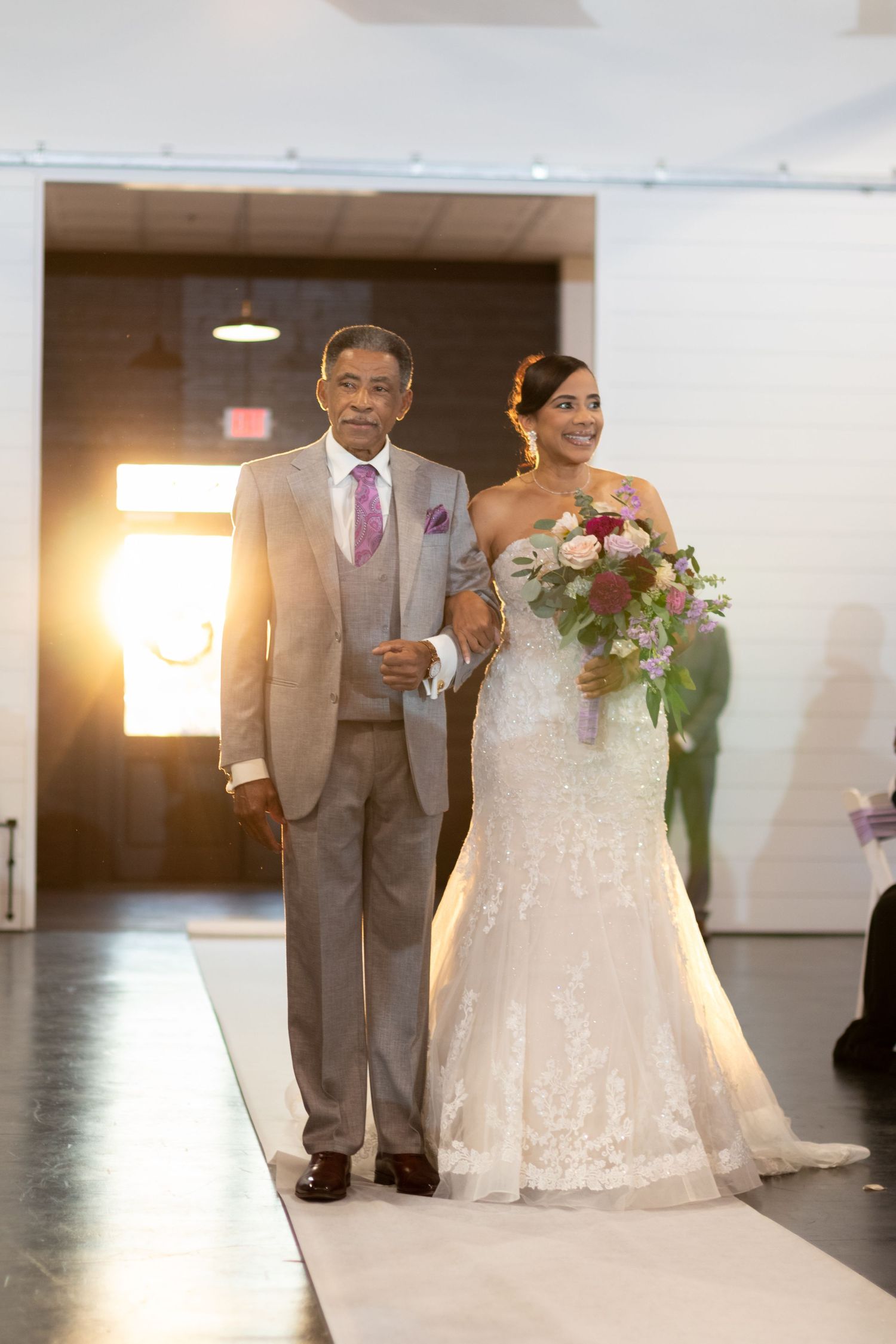 Two people walk arm-in-arm down a white aisle during a wedding ceremony, with bright backlighting creating a warm glow.