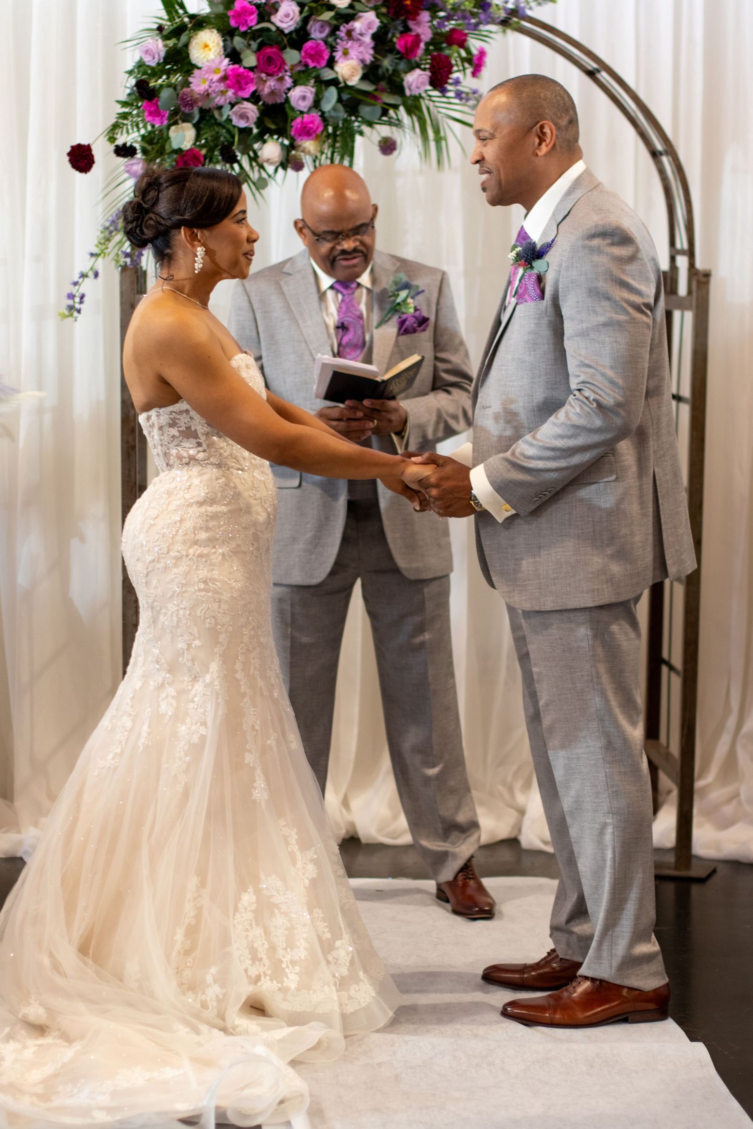 Wedding ceremony takes place under floral arch with couple in formal attire and officiant in gray suit.