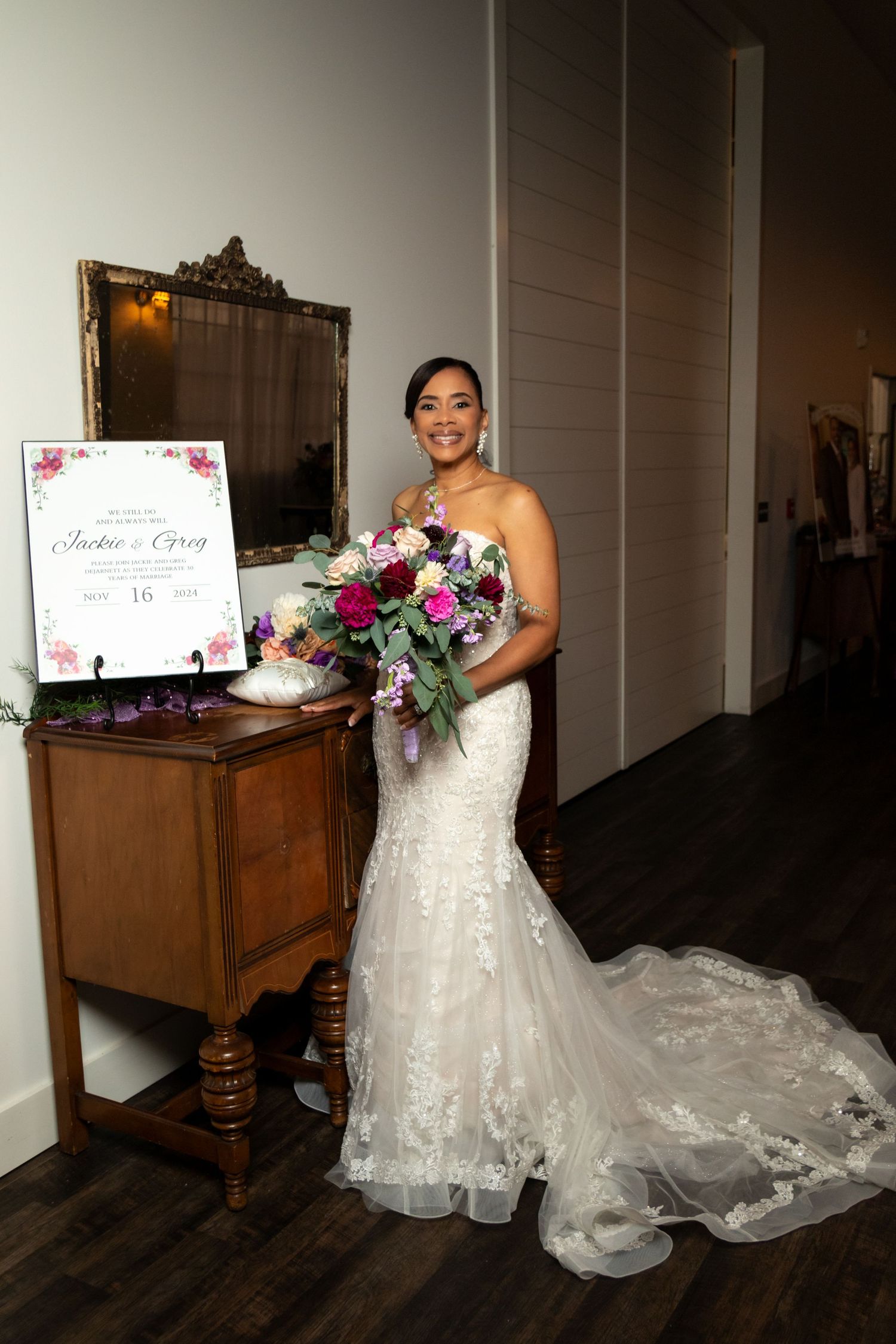 A bride in a flowing lace mermaid wedding gown holds a vibrant purple and pink bouquet next to an antique wooden table.