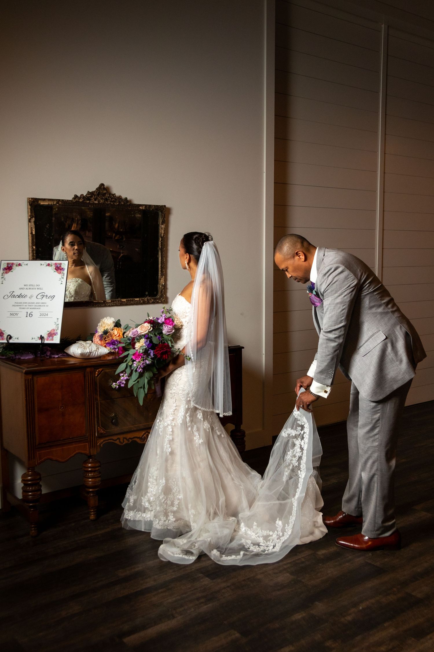 A tender moment captured as the groom helps adjust the bride's flowing lace wedding gown train before an antique mirror.