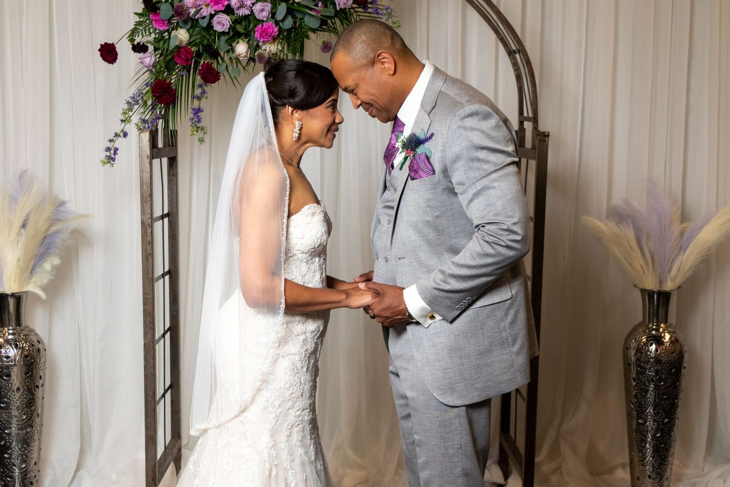 A couple shares an intimate moment during their wedding ceremony under a floral arch with pink blooms and elegant decorations.