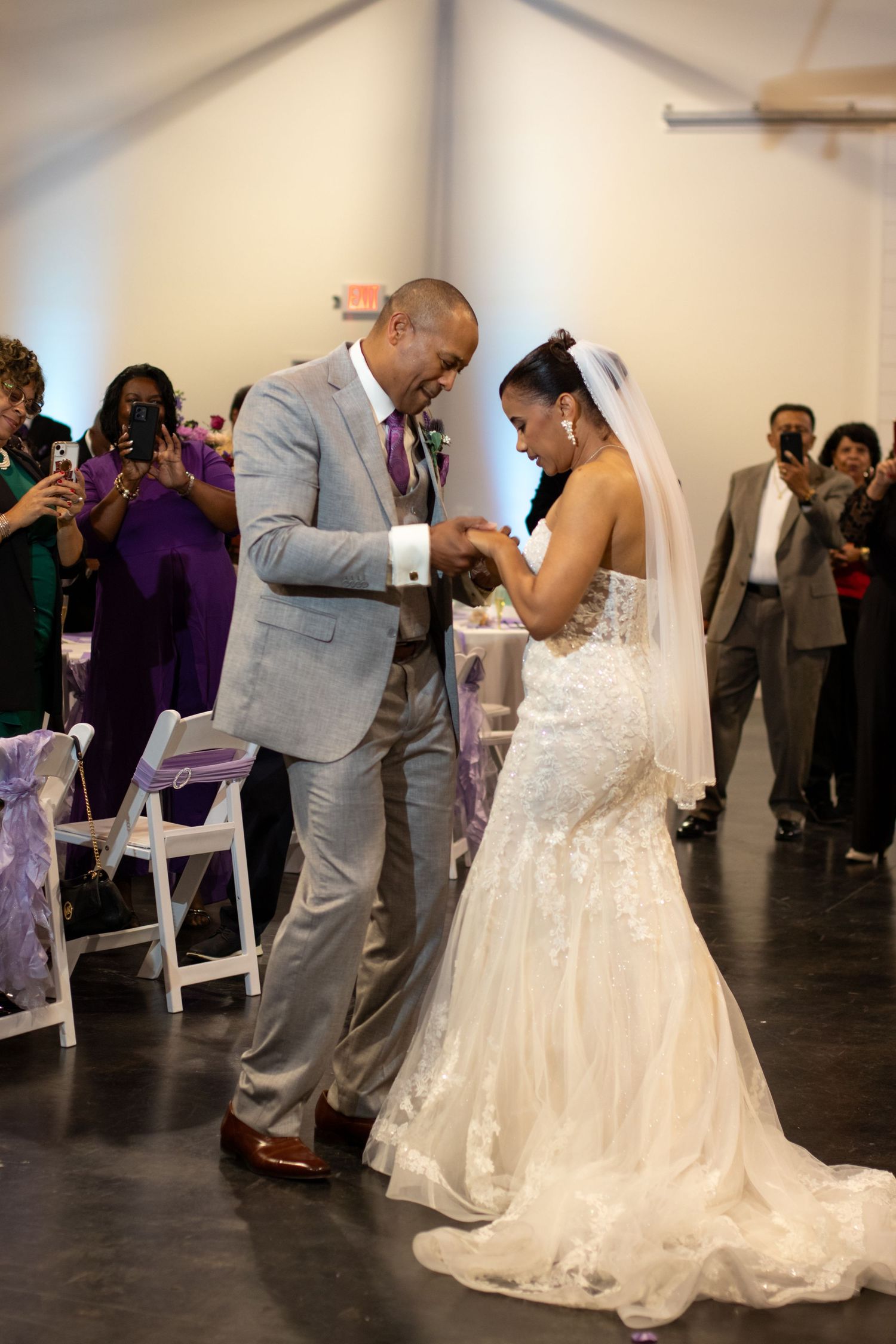 Newlyweds share their first dance together at reception venue with purple-dressed guests watching in background.