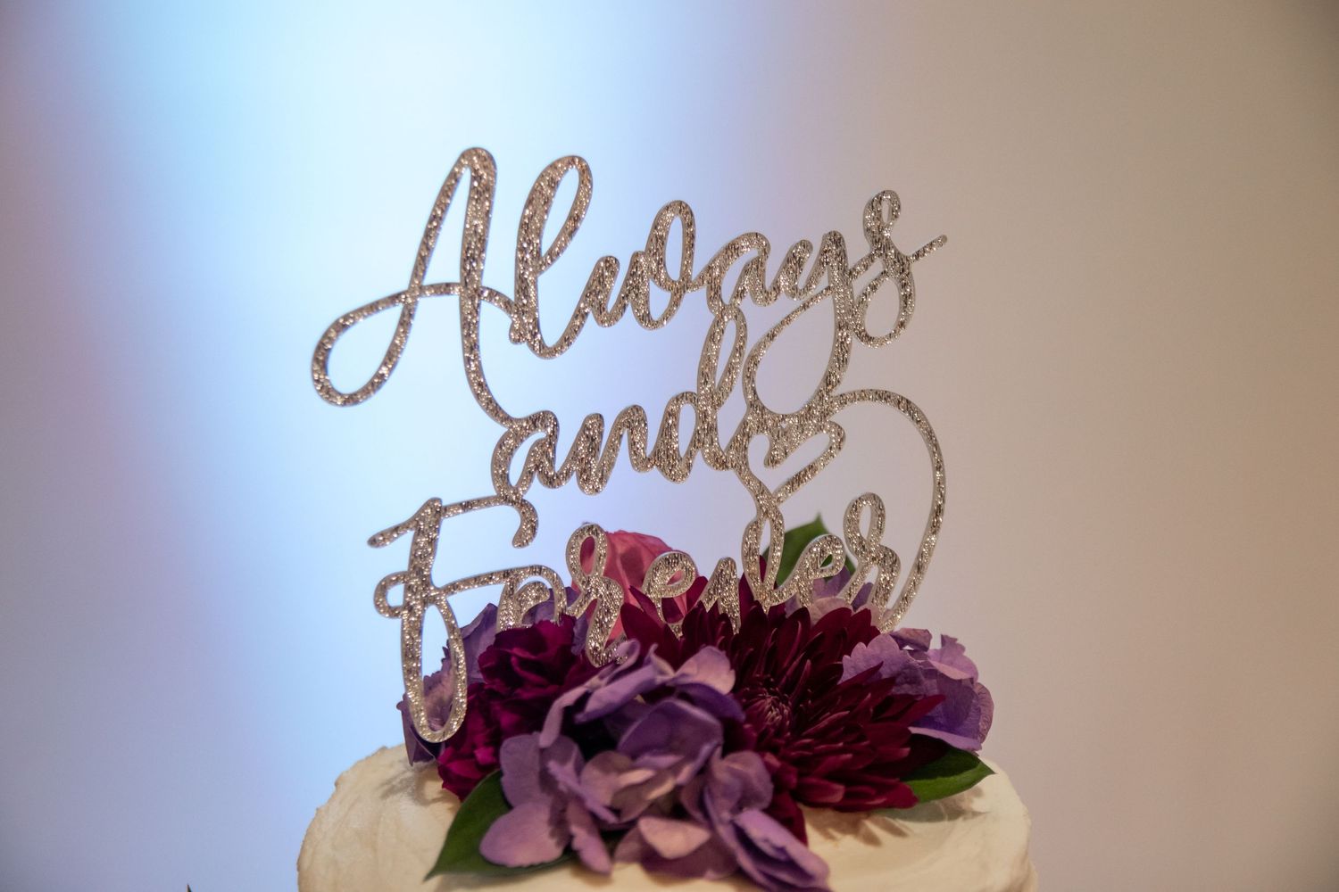 Gold glitter cake topper that reads 'Always and Forever' sits atop a white cake decorated with purple flowers.