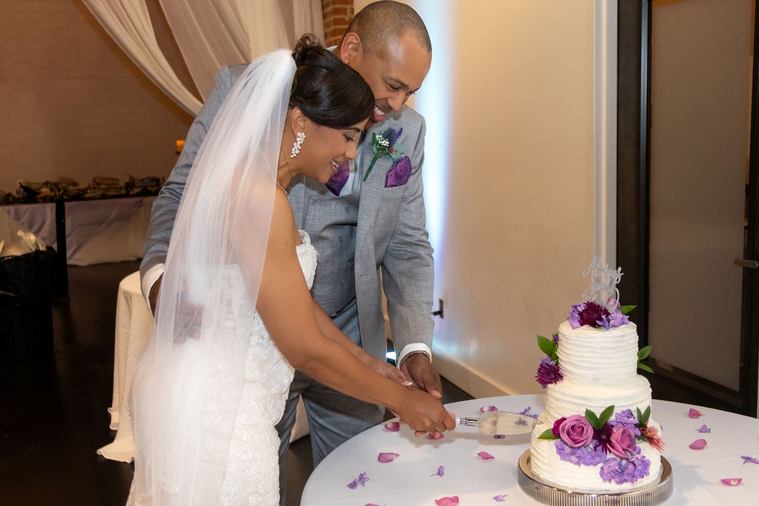 A couple in wedding attire cuts their white and purple decorated wedding cake together at the reception.