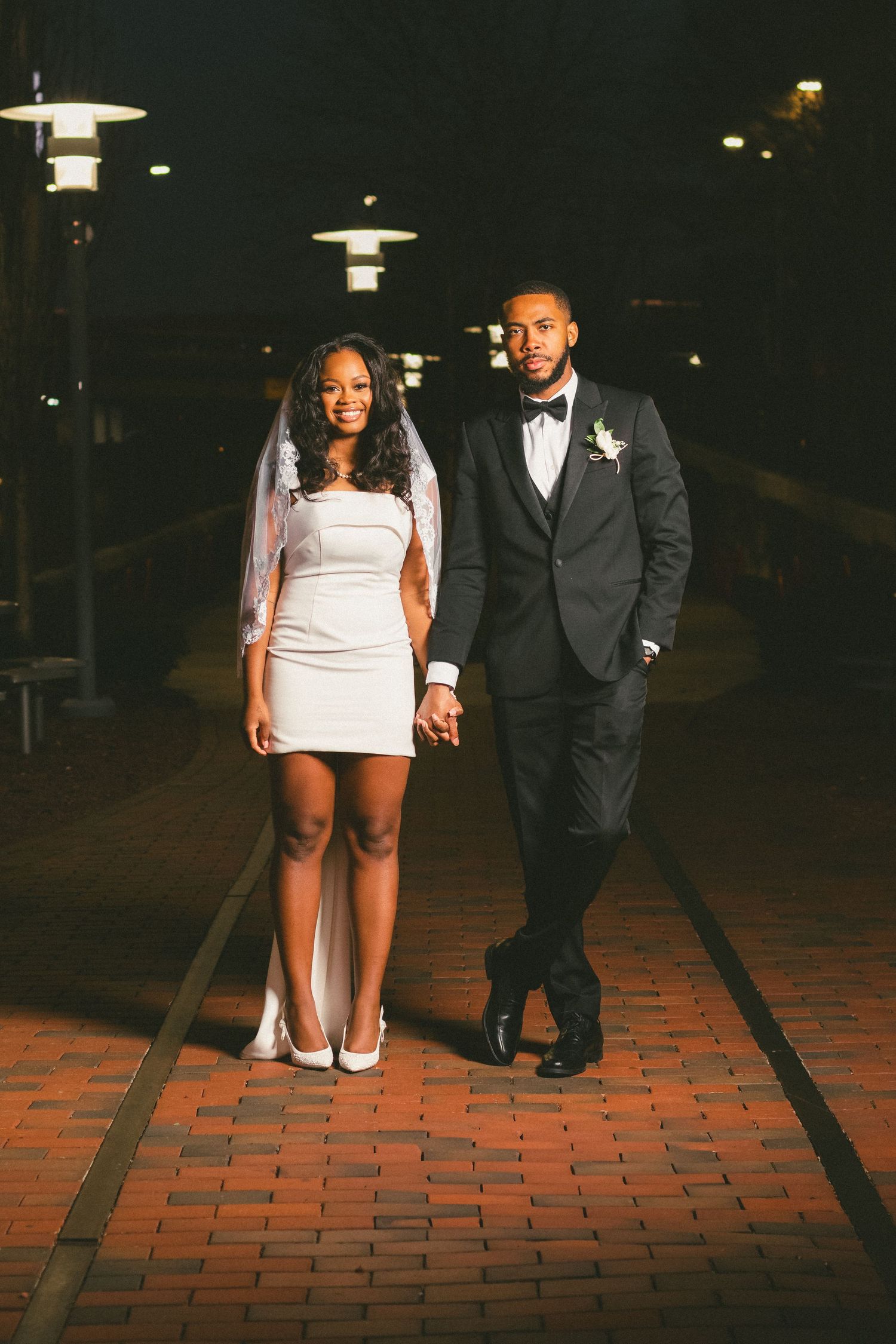 A couple poses at night on a brick pathway beneath vintage-style lamp posts during their formal event.