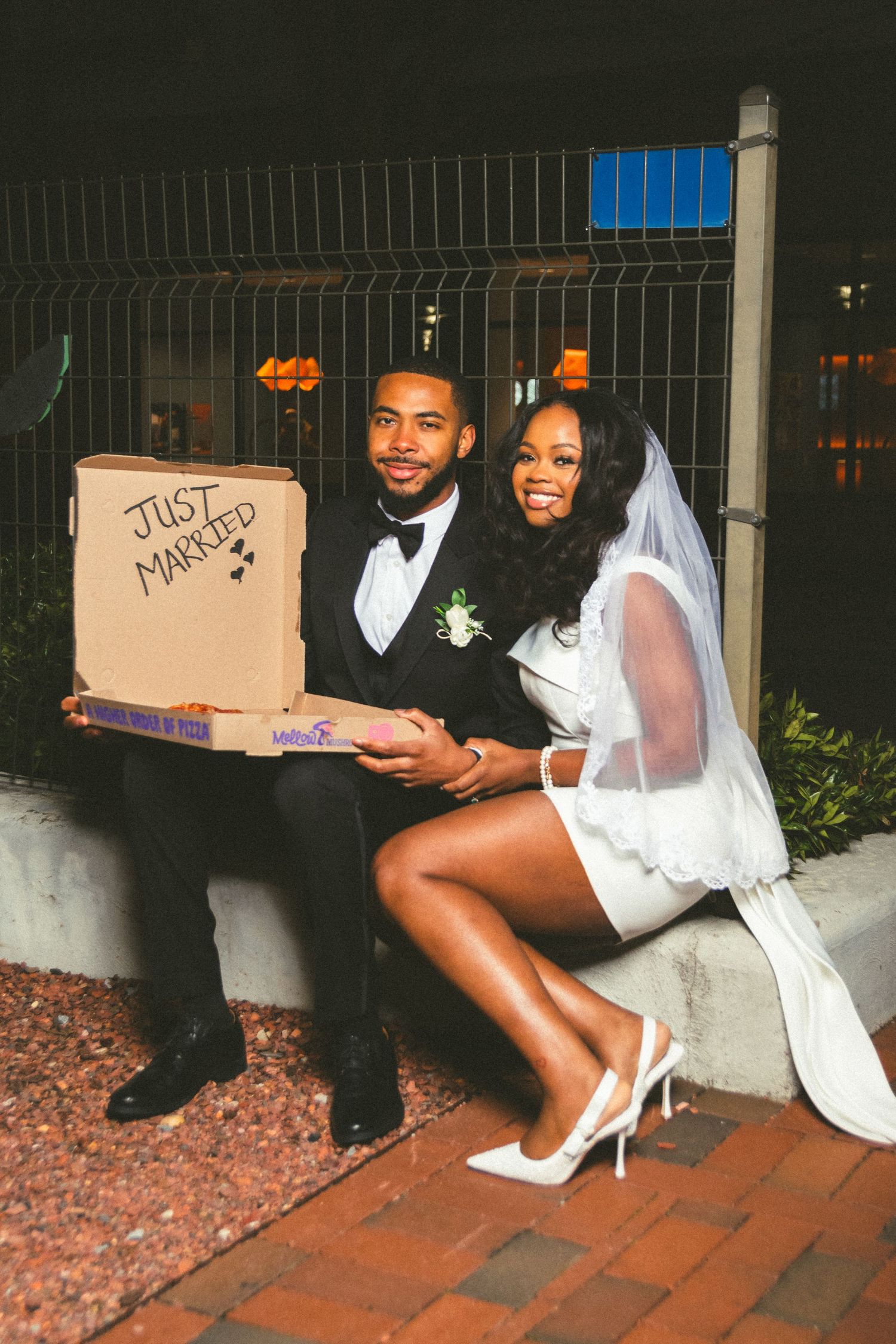 A couple in wedding attire poses with a cardboard box marked 'Just Married' on brick steps.