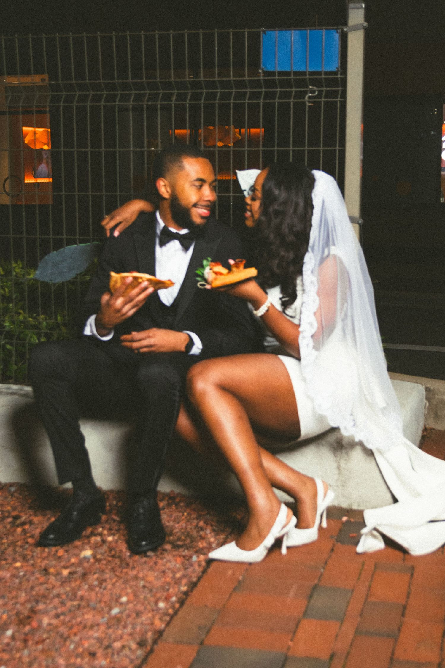 A couple in wedding attire share pizza while sitting on a brick patio at night.