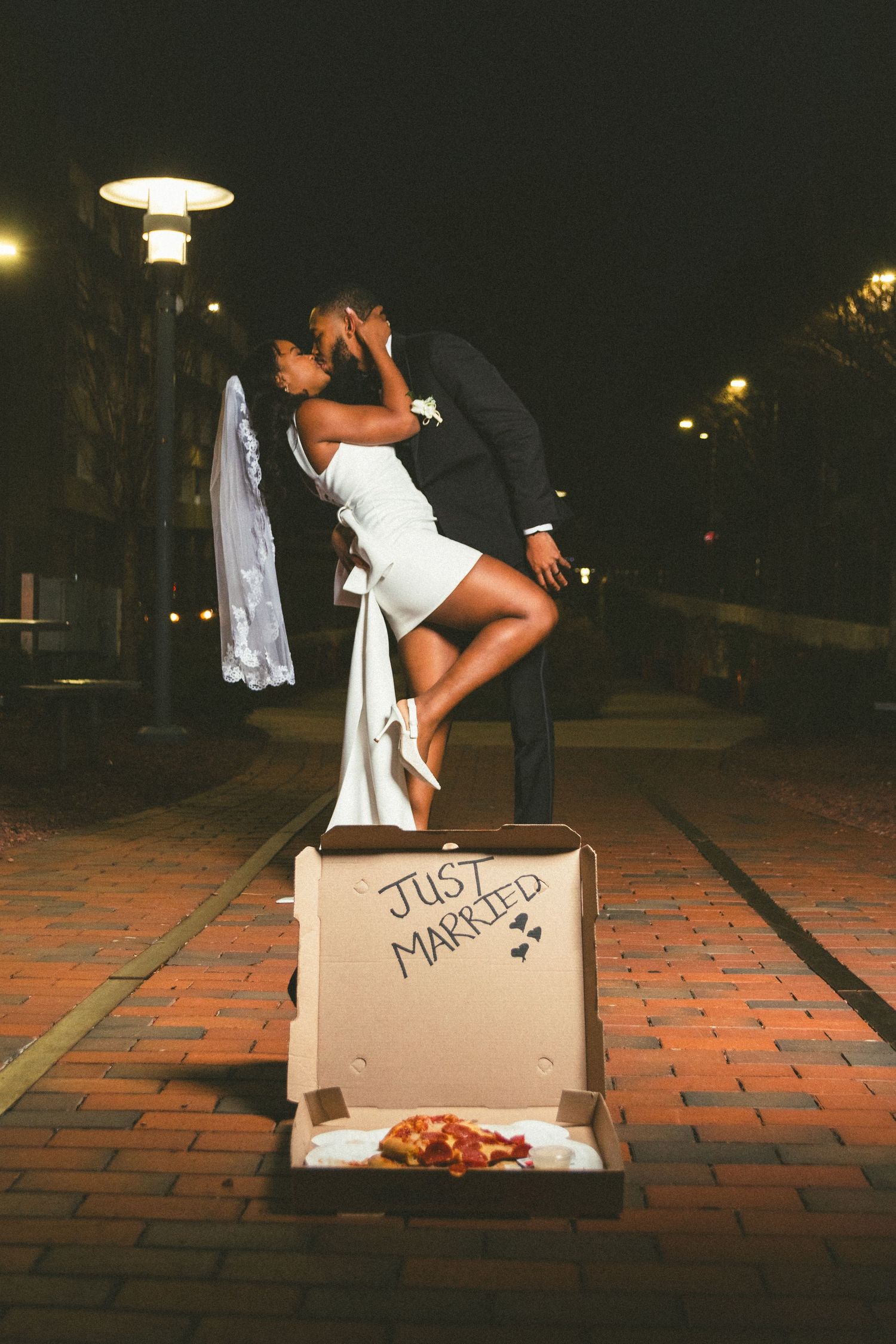 Romantic nighttime wedding photo with couple standing on pizza box marked 'Just Married' on brick sidewalk under street lights.