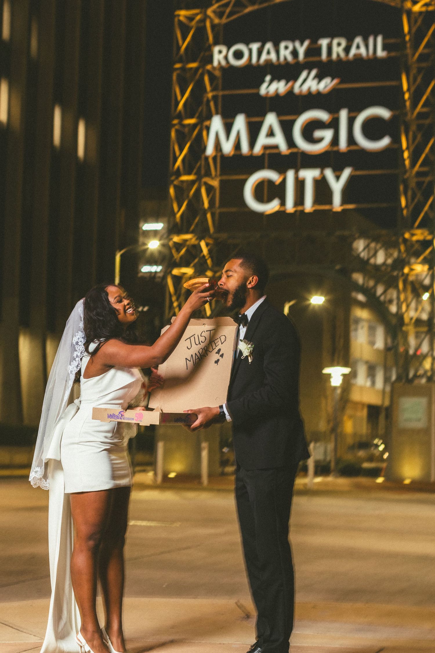 A couple celebrates their wedding at night in front of the illuminated Magic City Rotary Trail sign.
