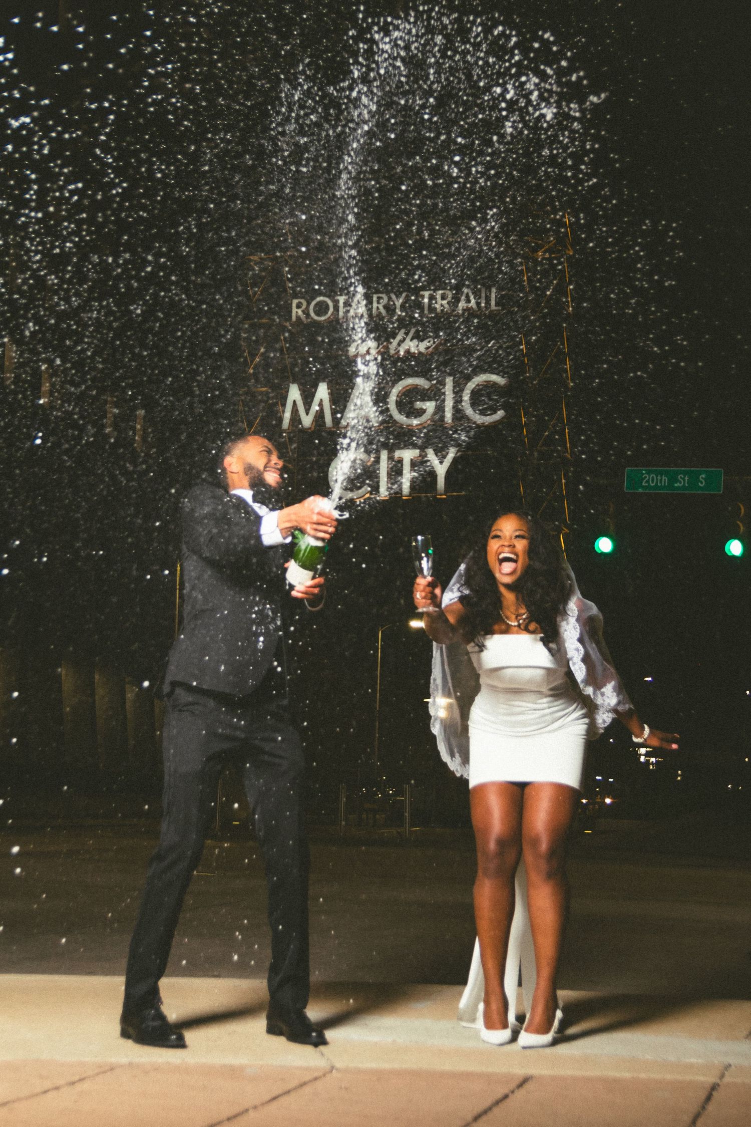 Celebratory couple popping champagne bottles at night with sparkling lights under a Magic City sign.
