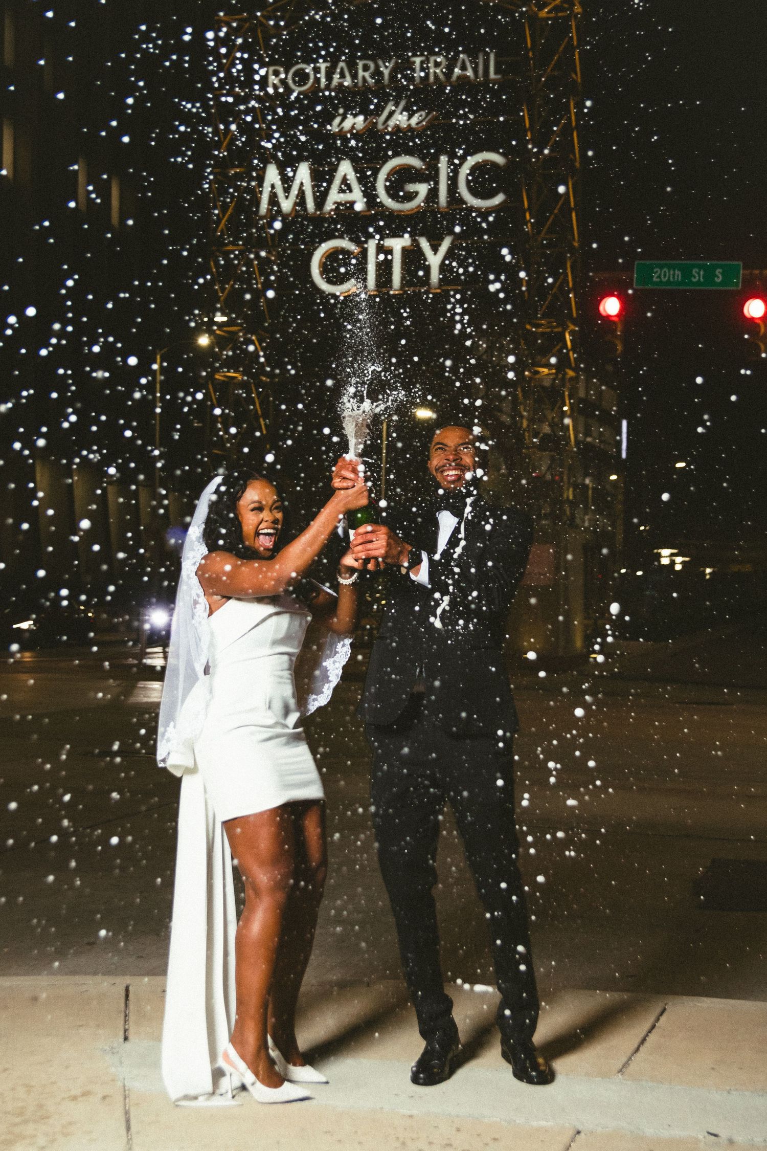 A couple pops champagne under a Magic City sign at night while being showered with glittering confetti.