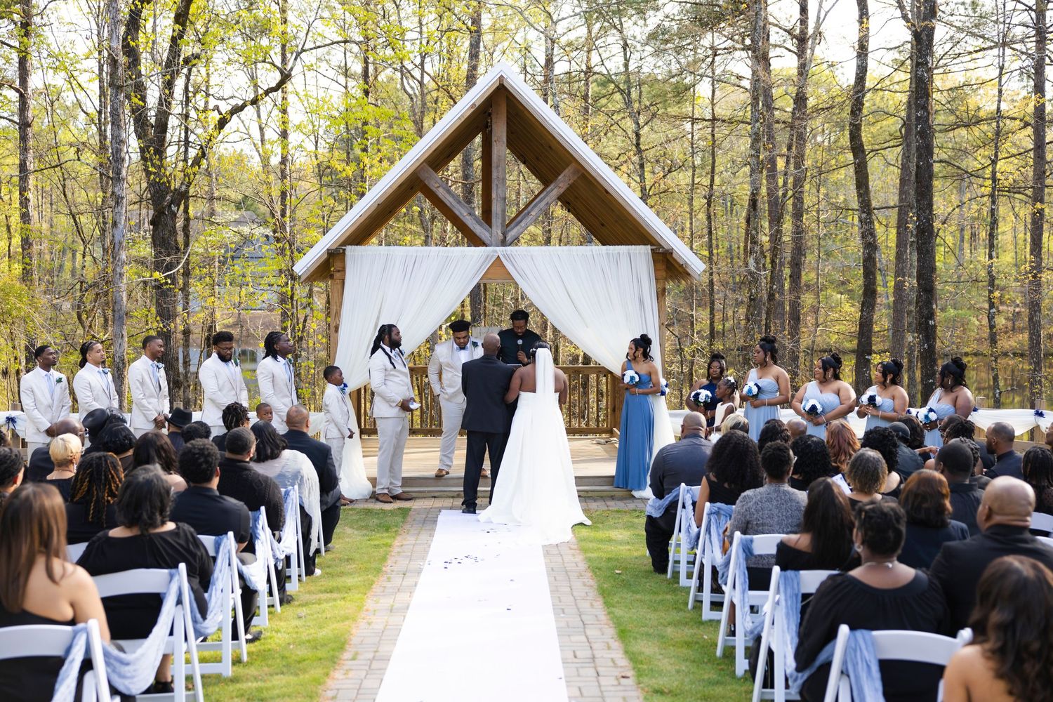 Outdoor wedding ceremony at a rustic wooden chapel surrounded by tall trees.