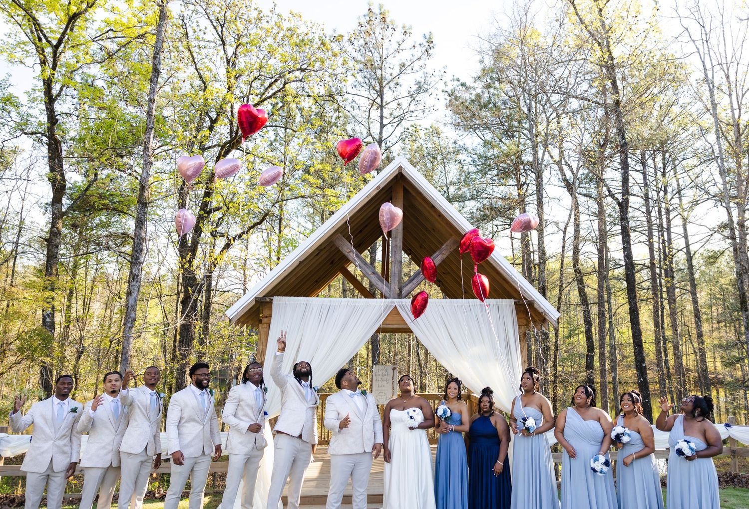 Wedding party in white attire posing with red balloons at a rustic outdoor chapel.
