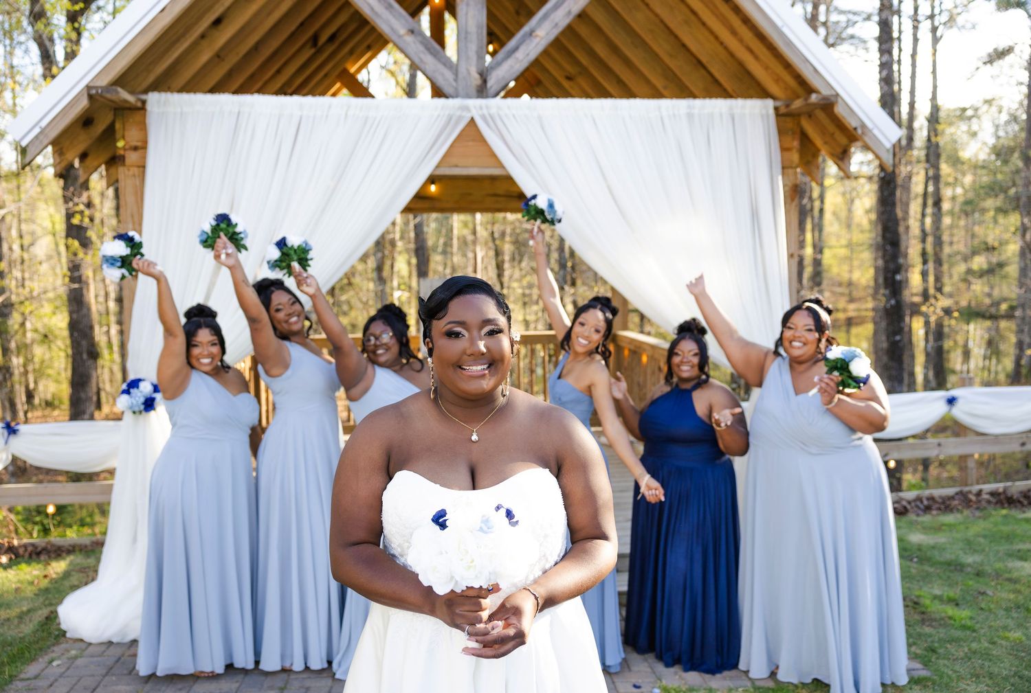 Bride and bridesmaids in light blue dresses pose together at rustic outdoor wedding venue.