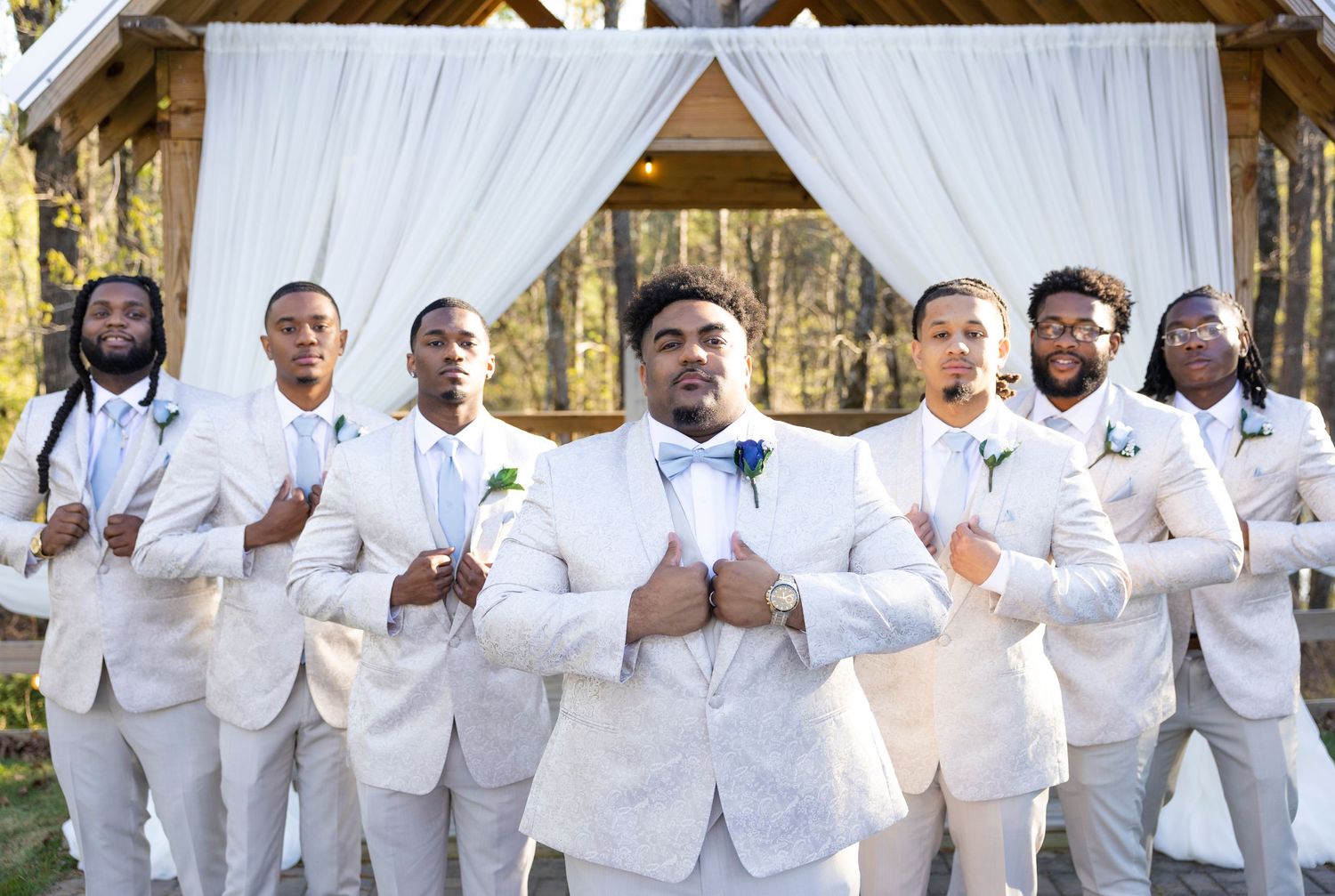 Groomsmen in crisp white suits pose together outdoors at a wedding venue.