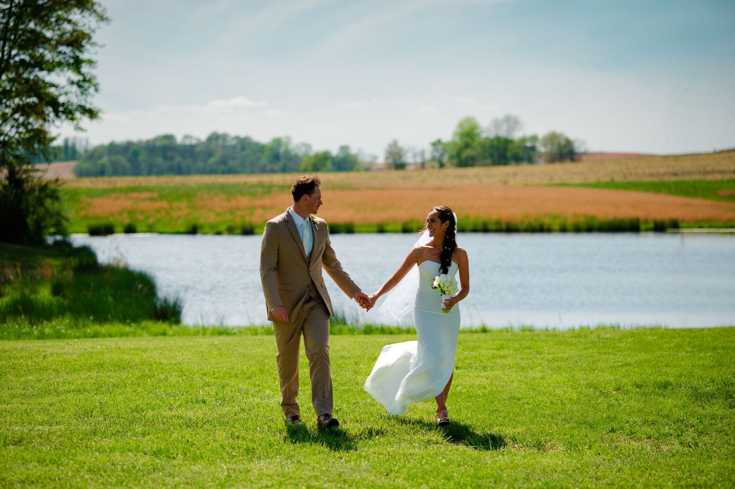 A couple in wedding attire holds hands on a grassy hill overlooking a scenic pond and autumn fields.
