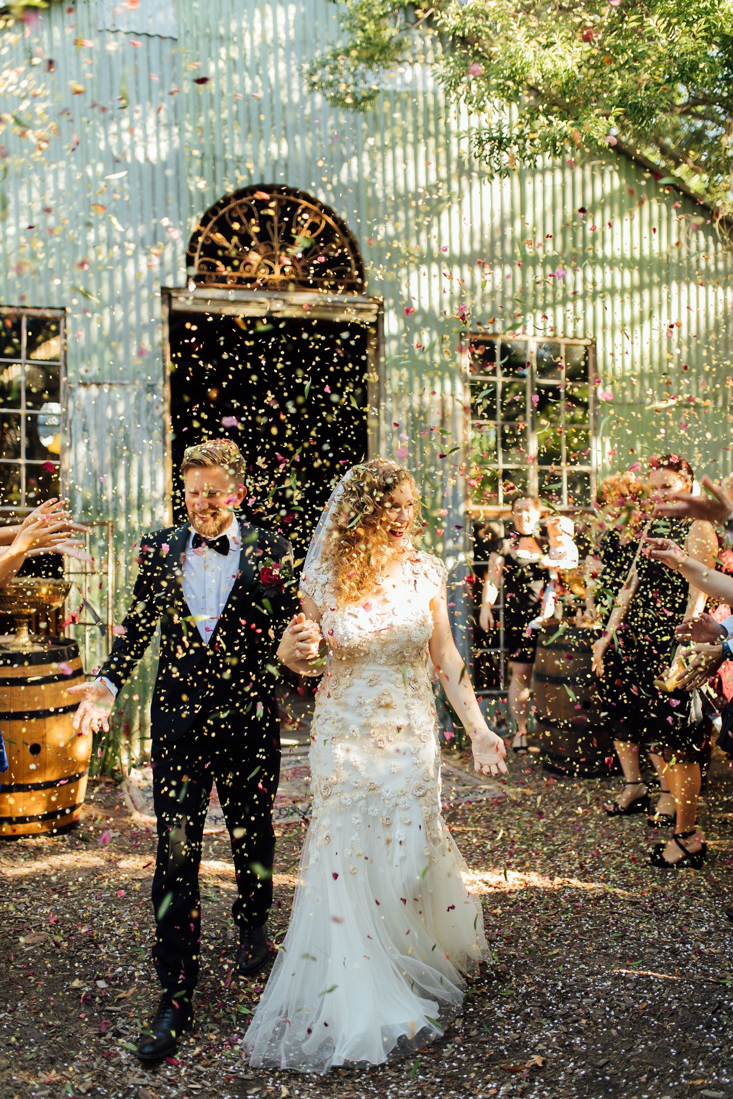 Newlyweds celebrate under a shower of confetti at a rustic outdoor venue with sunlight streaming through trees.