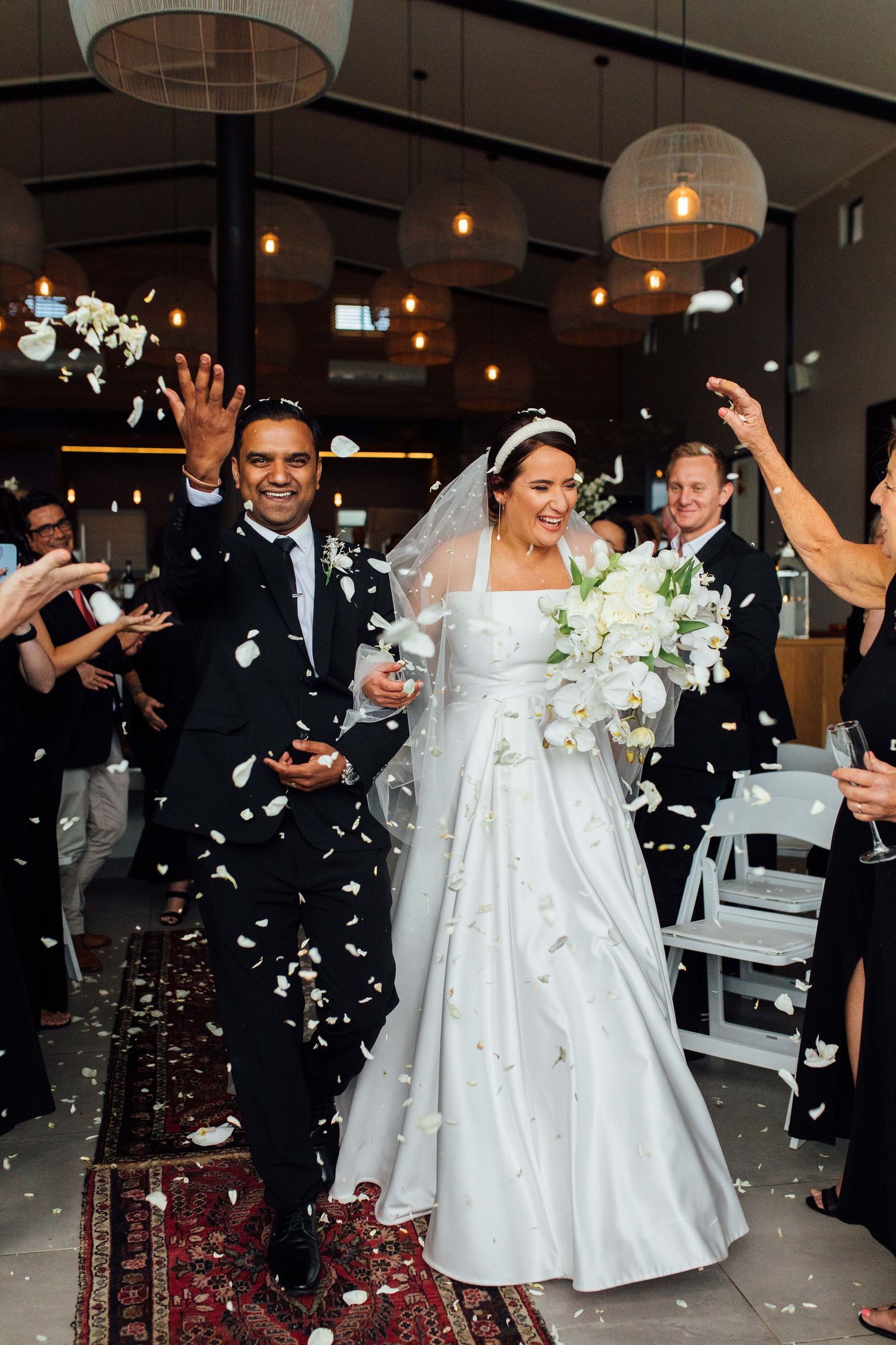Newlyweds celebrate with guests throwing white confetti in an indoor venue with warm lighting and elegant decor.