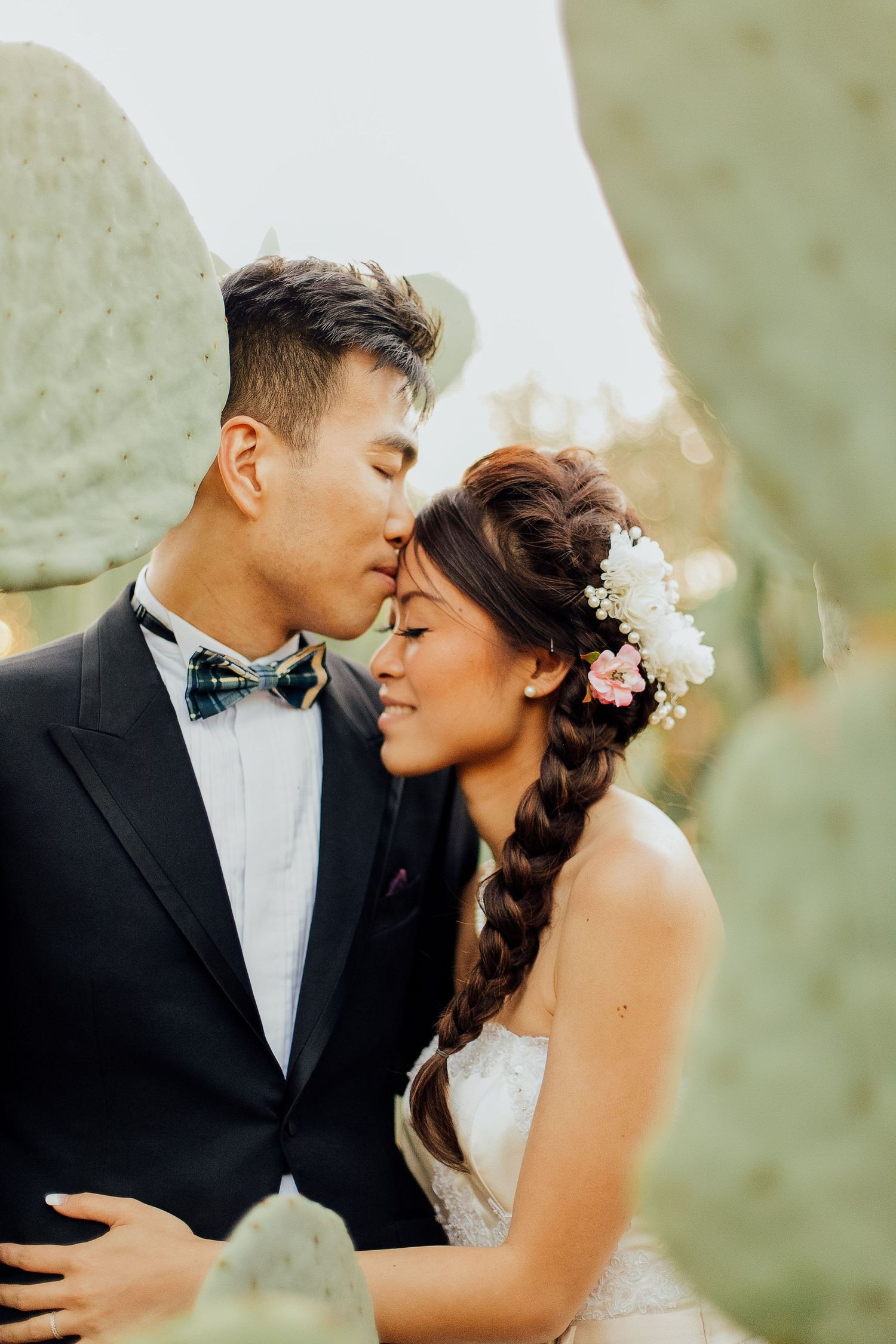 A romantic wedding portrait of a couple embracing among cactus plants, with the bride wearing floral hair accessories.