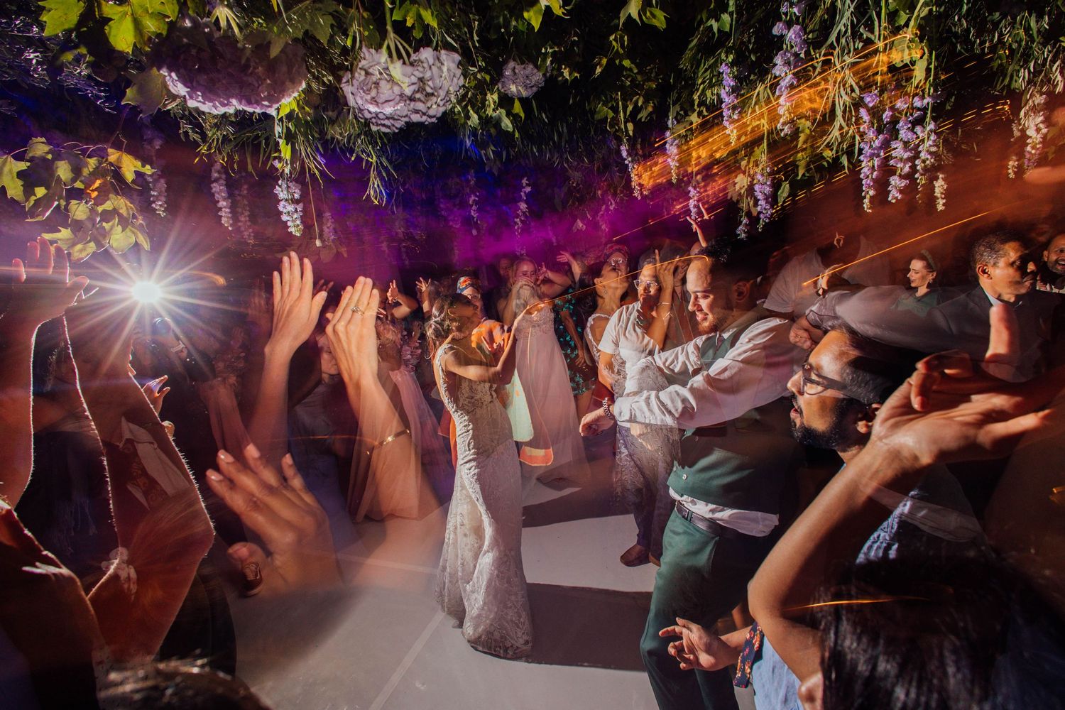 Party guests dance together on a lively dance floor illuminated by colorful lights and hanging floral decorations.