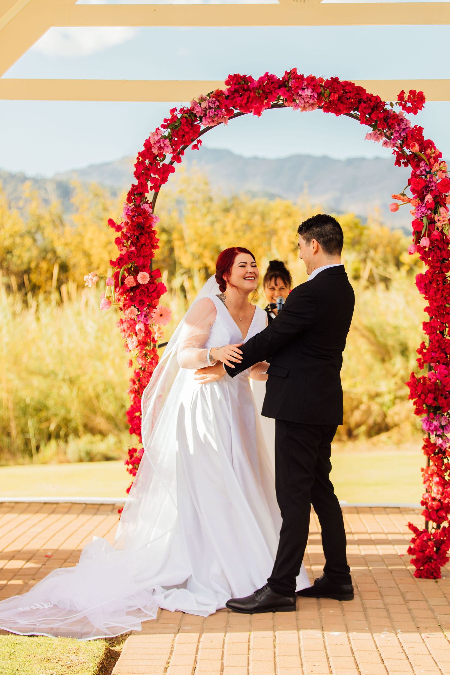 A wedding ceremony takes place under a vibrant pink floral arch against a golden desert landscape background.